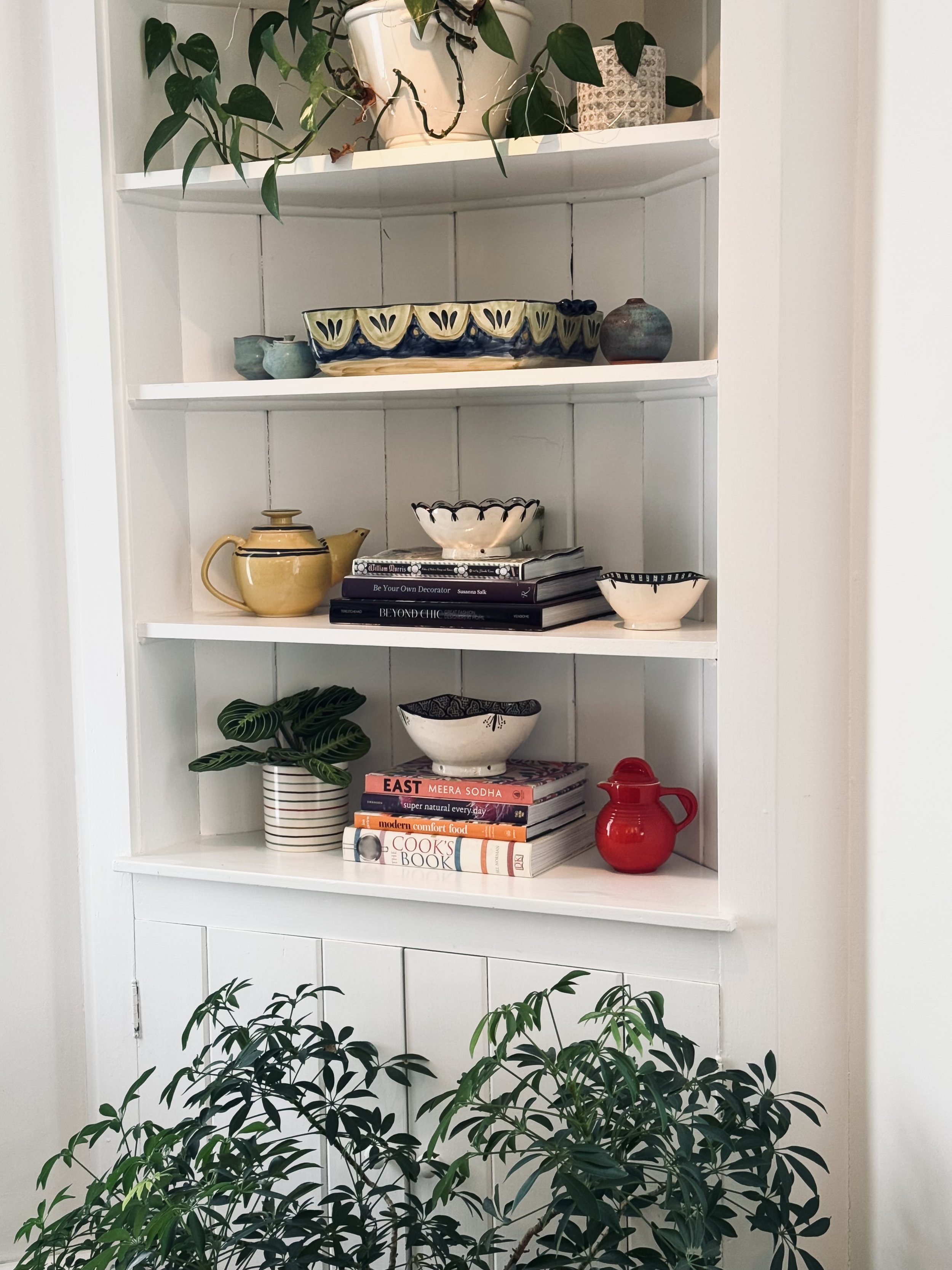 White shelf displaying potted plants, ceramic bowls, teapot, books, and red pitcher