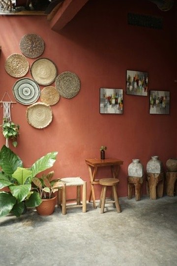 Cozy patio corner with a small wooden table and stool, surrounded by large potted plants. Woven baskets form a decorative arrangement on the brown wall alongside abstract paintings. Rustic clay pots line up below the paintings.