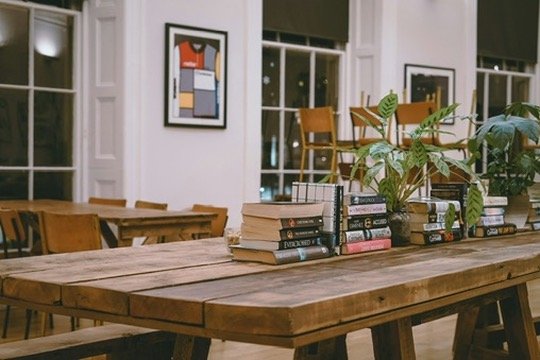 Cozy café interior with wooden tables, stacked books, and potted plants. Modern art on white walls, soft lighting, chairs, and large windows present.