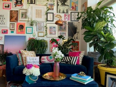 Eclectic home interior with a dark blue sofa, colorful throw pillows, and potted plants. A gallery wall displays an array of framed art and photos. A coffee table holds vases with white flowers and stacked books.