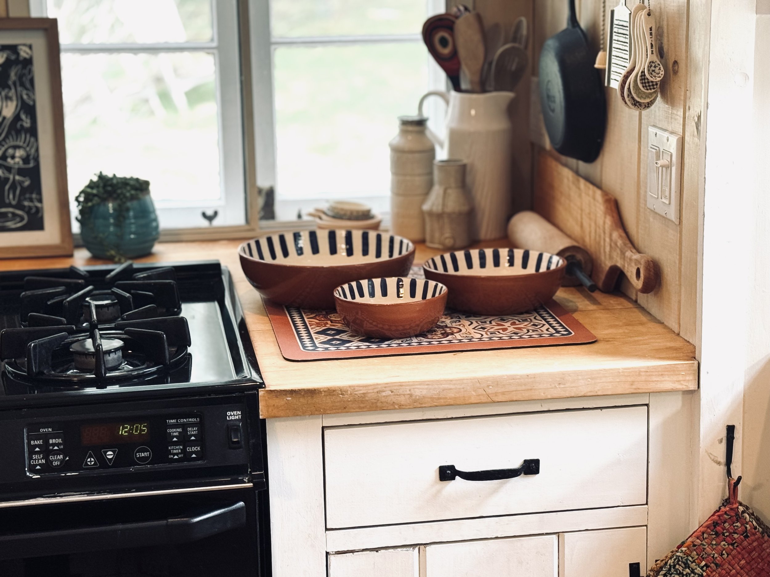 Rustic kitchen with wooden countertops, three brown bowls with black stripes, a gas stove, cookware, cutting boards, and measuring spoons hanging on the wall.