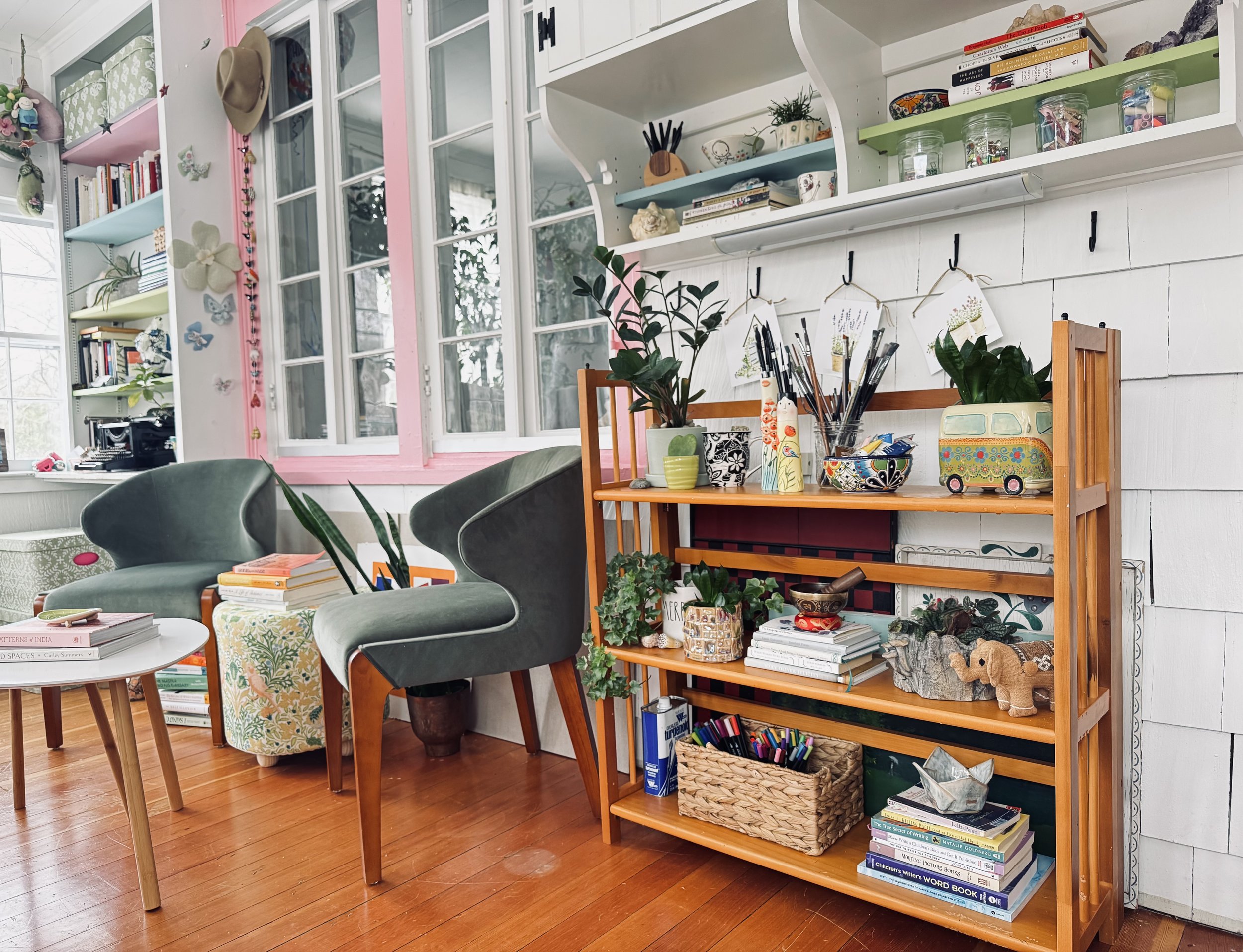 Cozy room interior with two green chairs, wooden shelving with plants and art supplies, books, and decorative items like pottery and a toy van. Shelves and books decorate the surrounding walls.