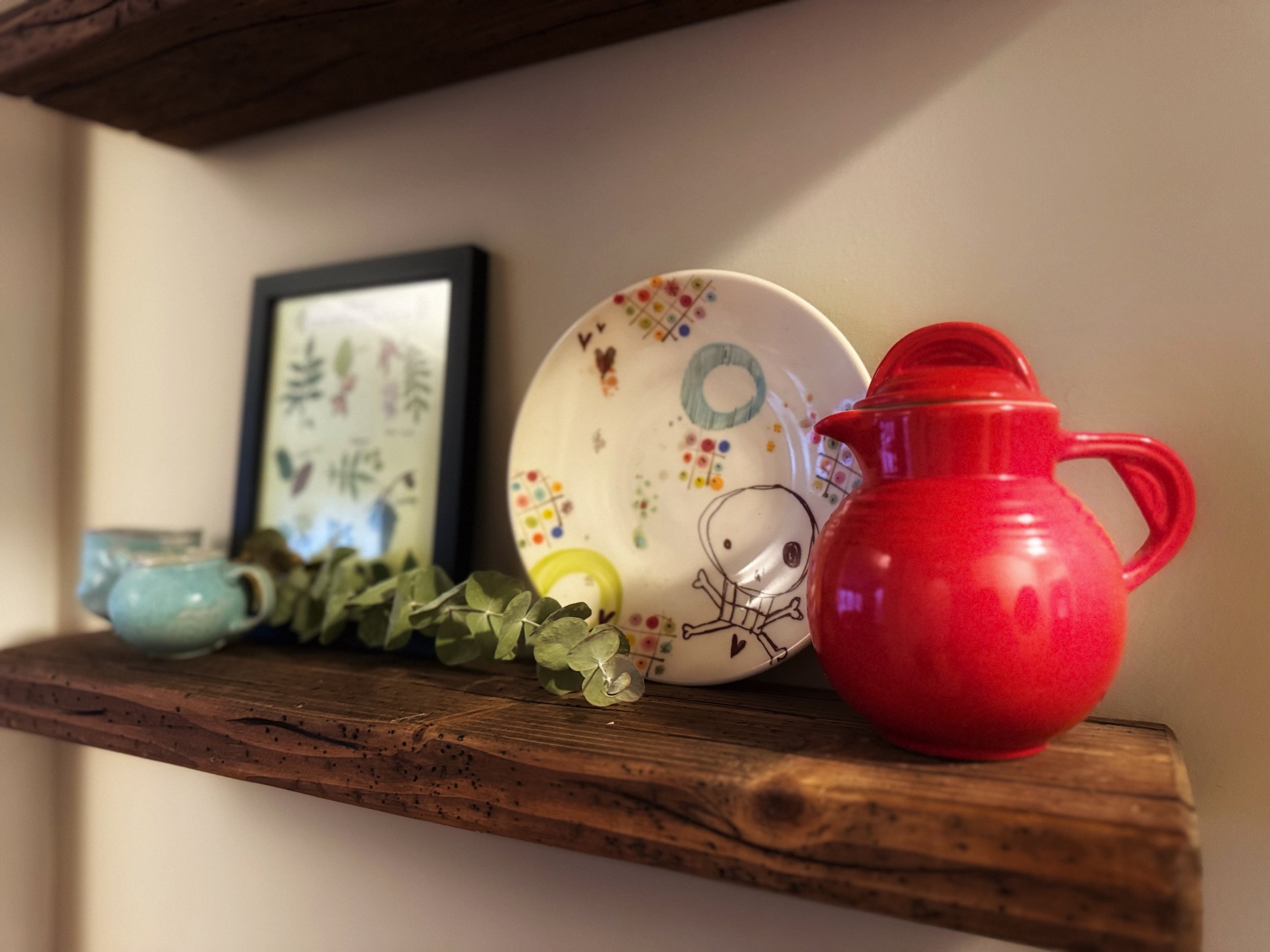 Decorative wooden shelf with a red pitcher, artistic plate with a cartoon character, framed botanical print, light blue ceramic cup, and eucalyptus leaves.