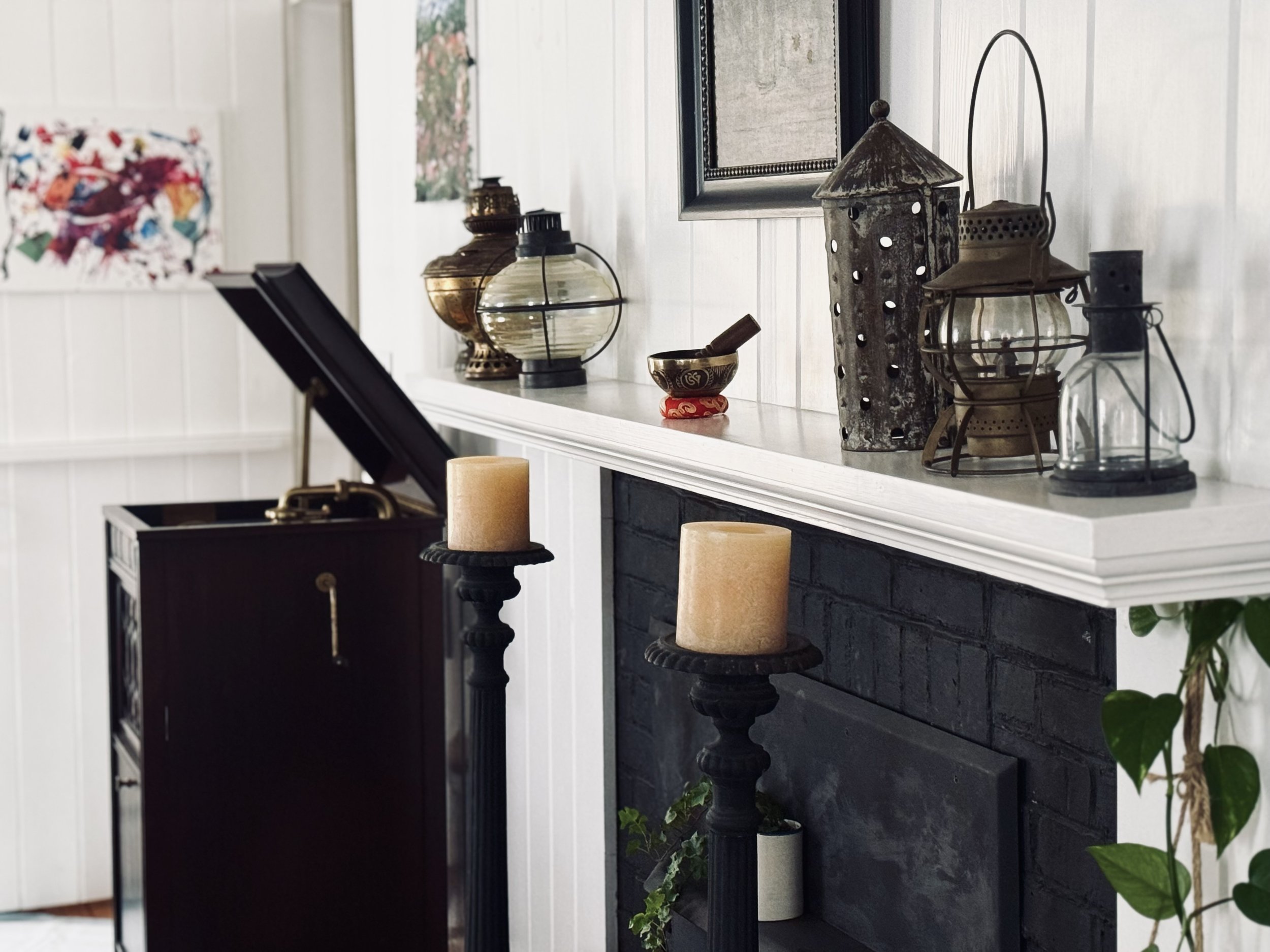 Vintage room interior with a gramophone, decorative artifacts on a white mantel, candles on black stands, and abstract paintings on white paneled walls.