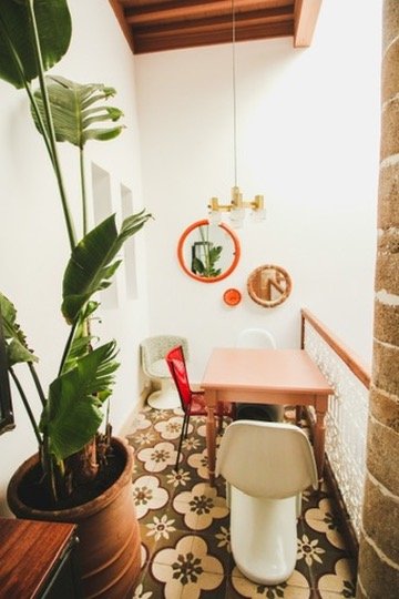 Small dining area with a pink table, white chairs, potted green plant, decorative mirrors, pendant light, and patterned floor tiles.