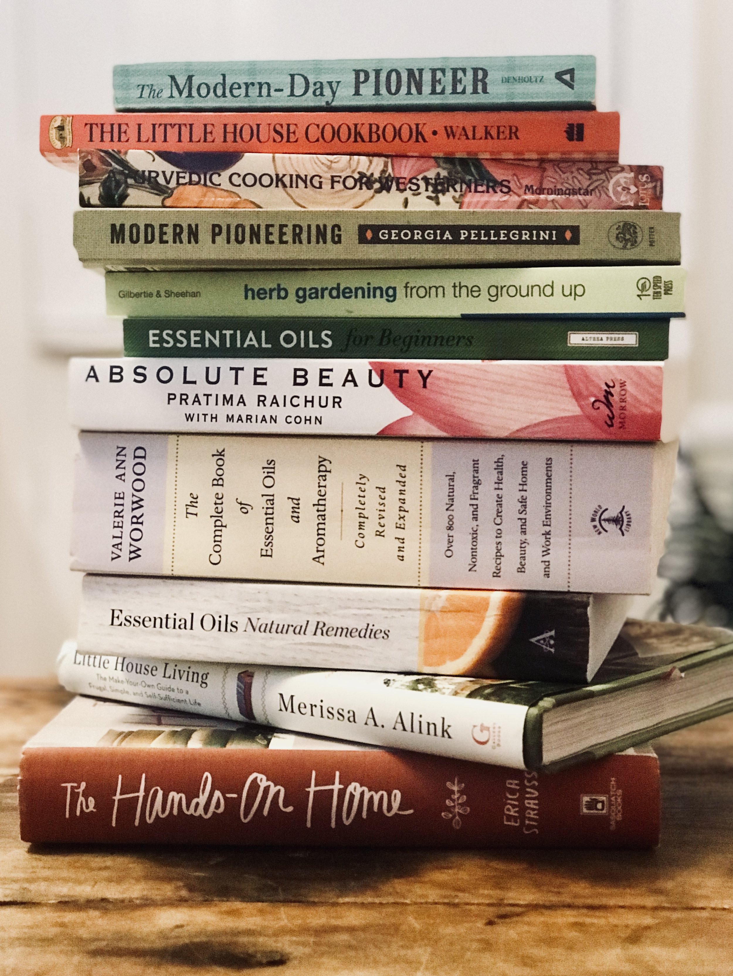 Stack of books on various topics including pioneering, cooking, gardening, essential oils, and home living, with titles like 'The Modern-Day Pioneer' and 'The Hands-On Home.'