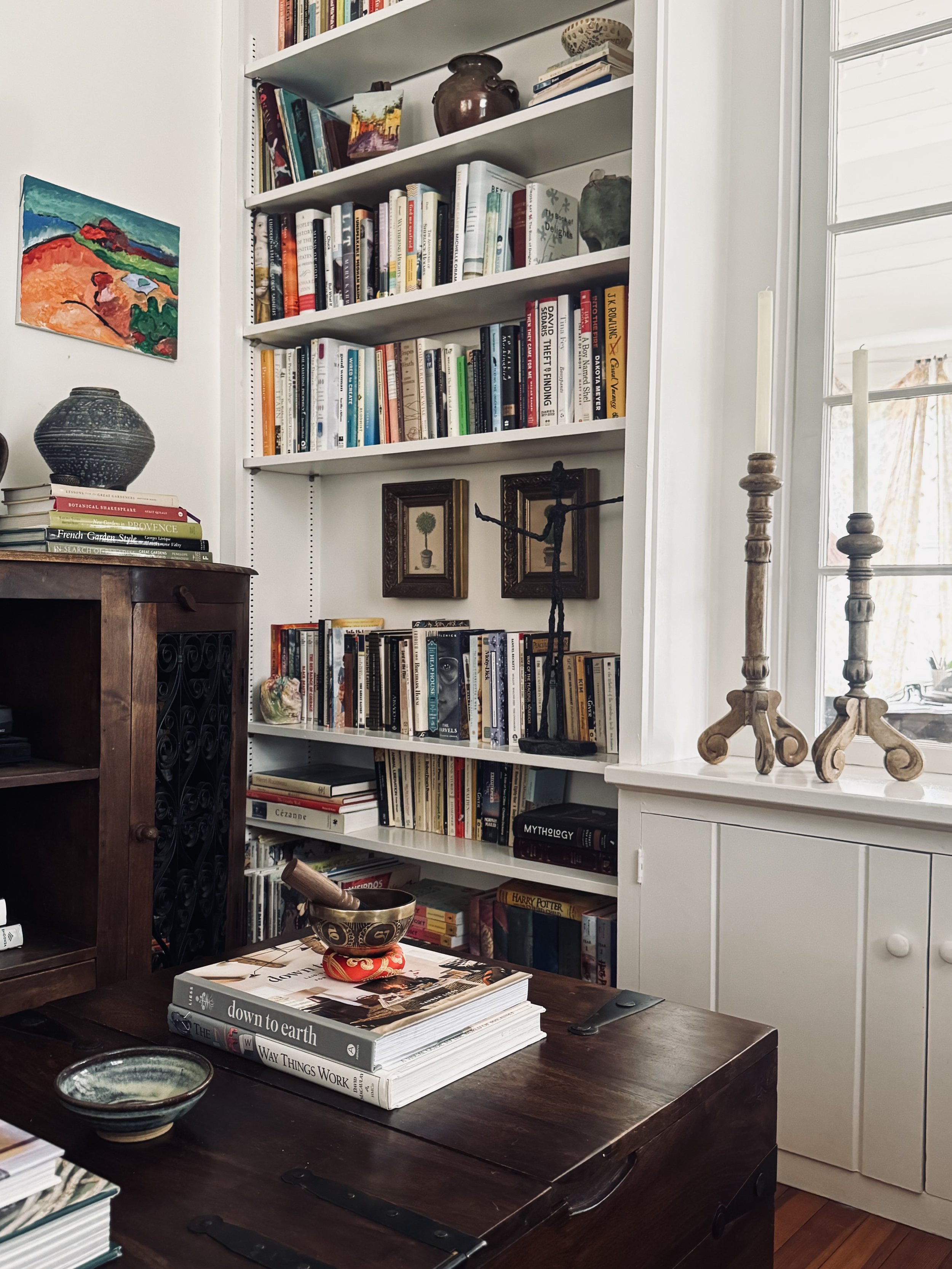 Cozy living room featuring a bookshelf filled with books, decorative items like a ceramic pot and a small painting, and a wooden coffee table with a mortar and pestle. Two tall candlesticks and a decorative sculpture are near a window.