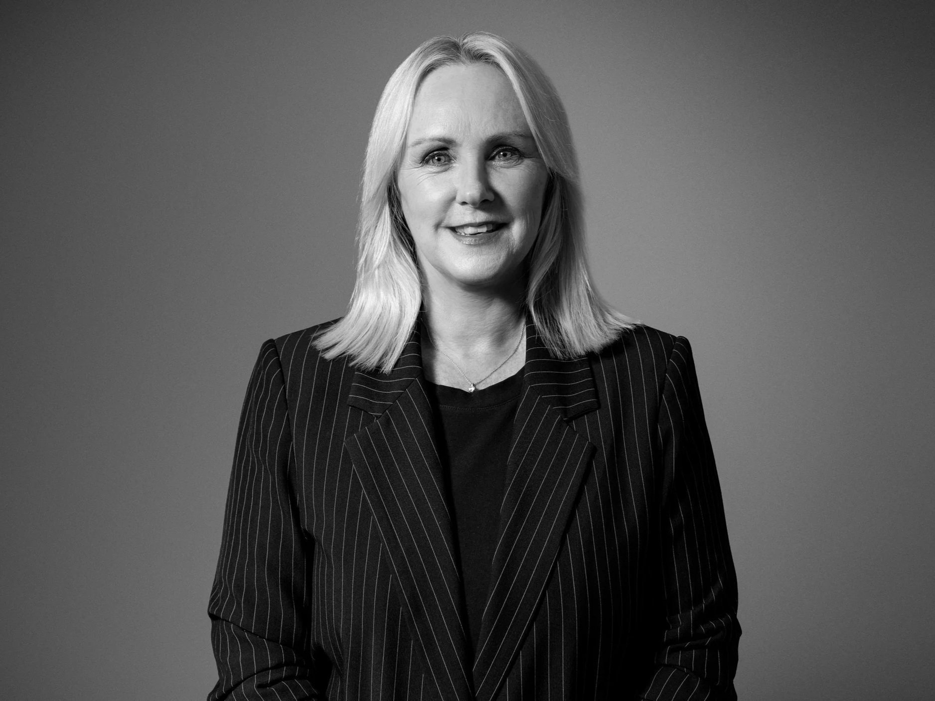 Black and white portrait of a woman with shoulder-length light hair, wearing a pinstripe blazer and a necklace, smiling softly, against a plain background.