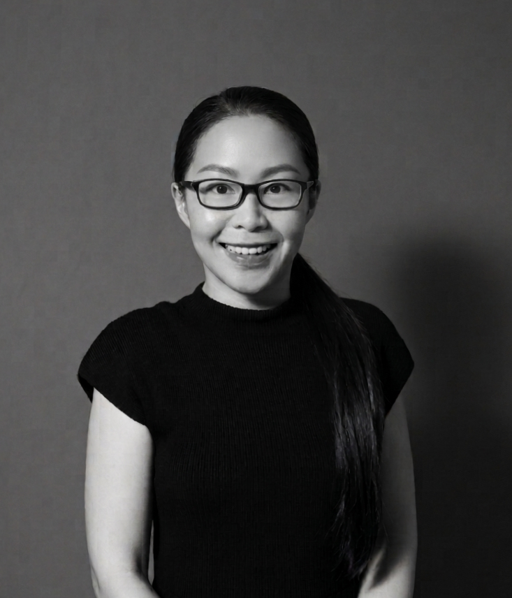 A woman with long dark hair and glasses smiling, wearing a black short-sleeve top, standing against a plain background.