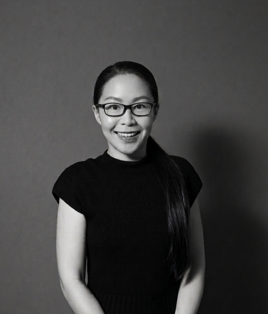 A smiling woman with glasses and long dark hair in a black top, standing against a plain backdrop.