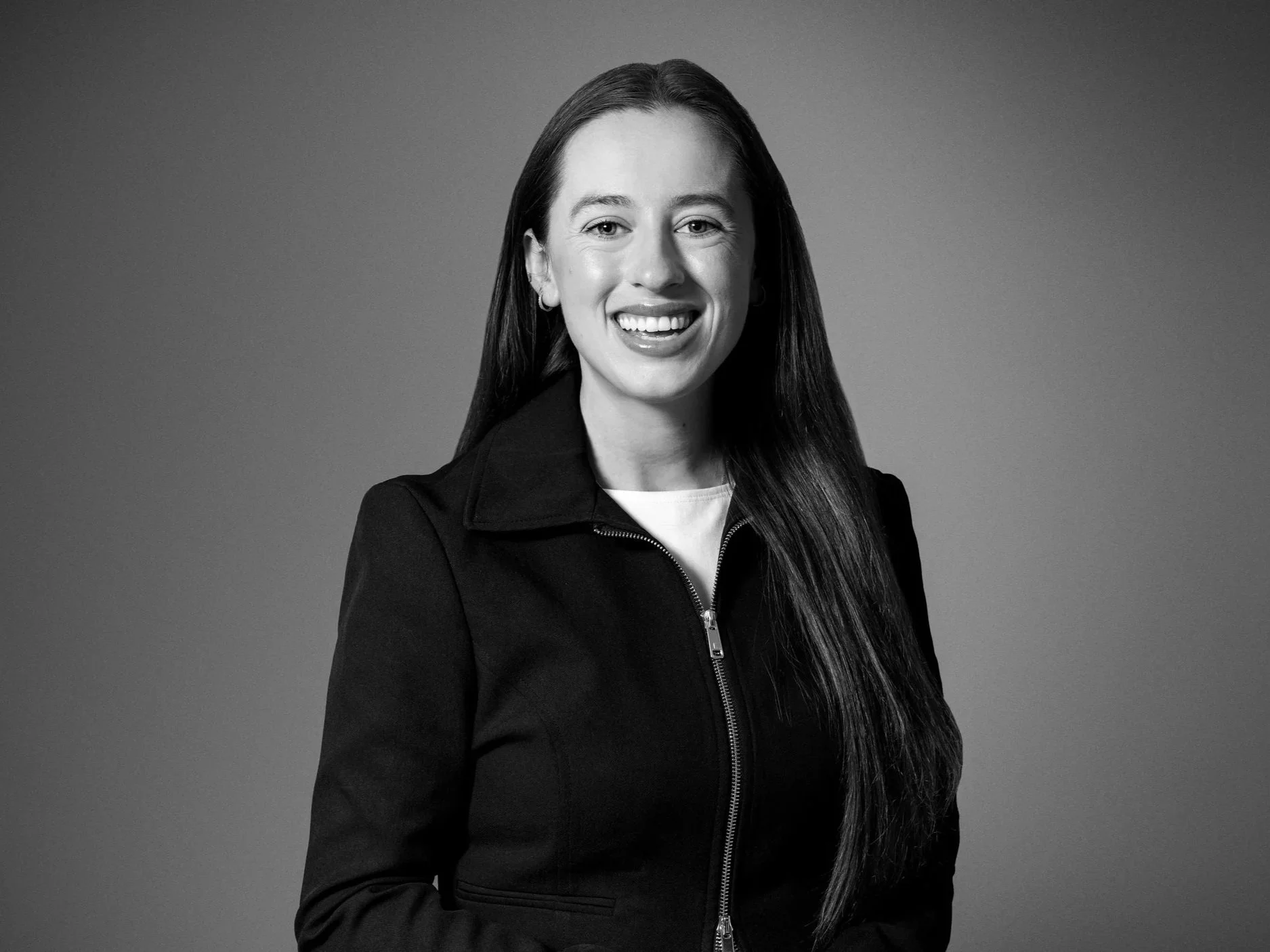 Black and white portrait of a woman smiling, wearing a dark jacket with a zipper, against a plain background.