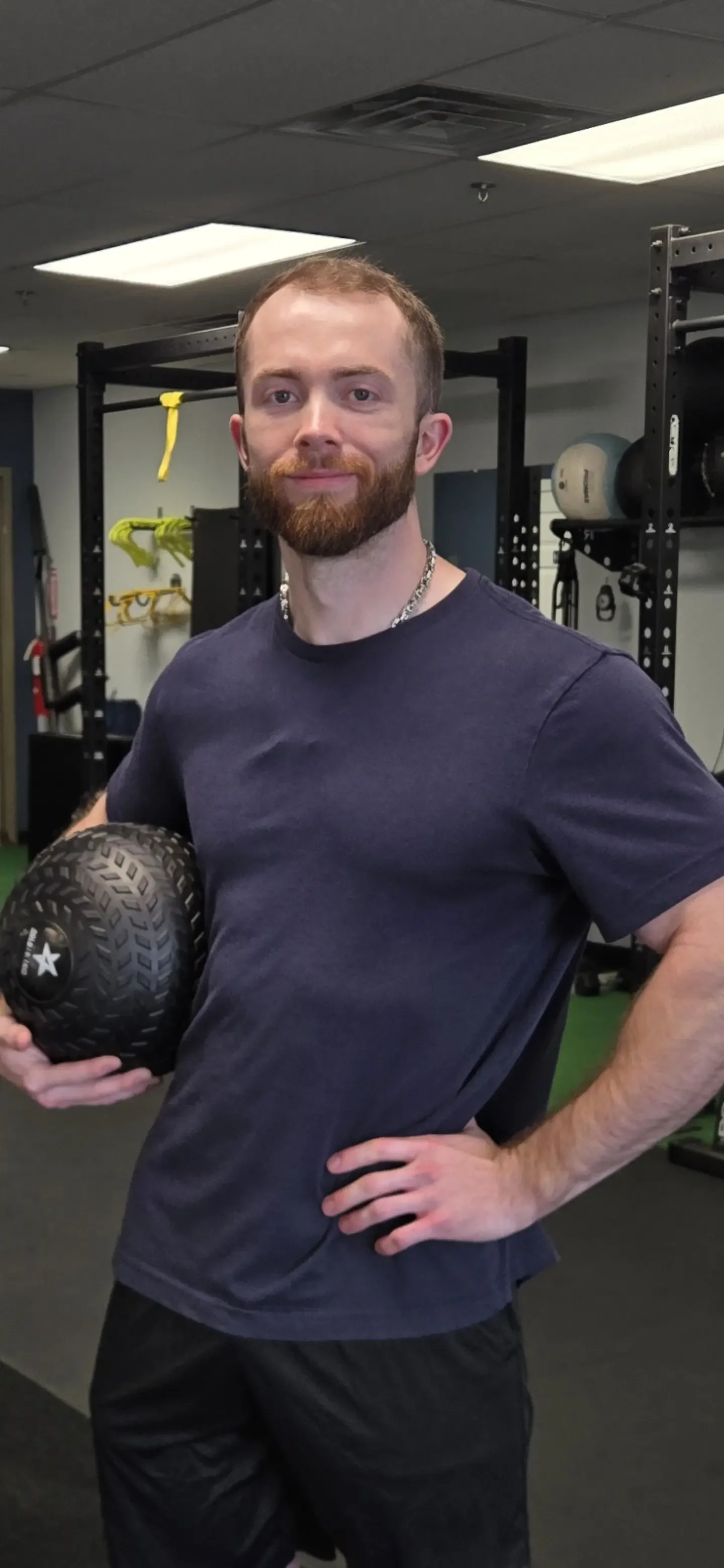 A man with a beard and short hair holding a black textured medicine ball in a gym, wearing a blue T-shirt and a chain necklace.