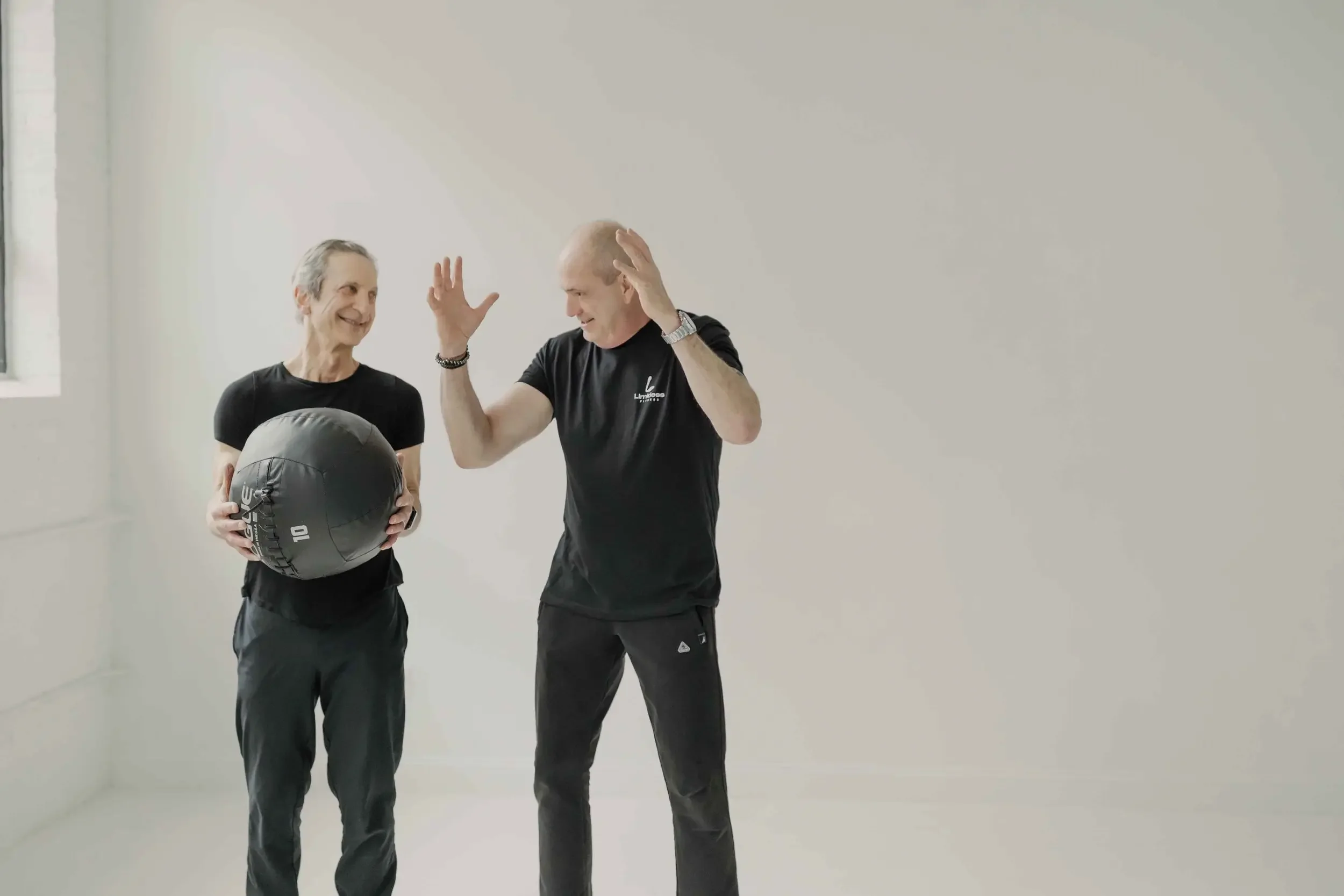 Two men in black workout attire, one holding a medicine ball, engaged in a friendly conversation in a brightly lit room with white walls.