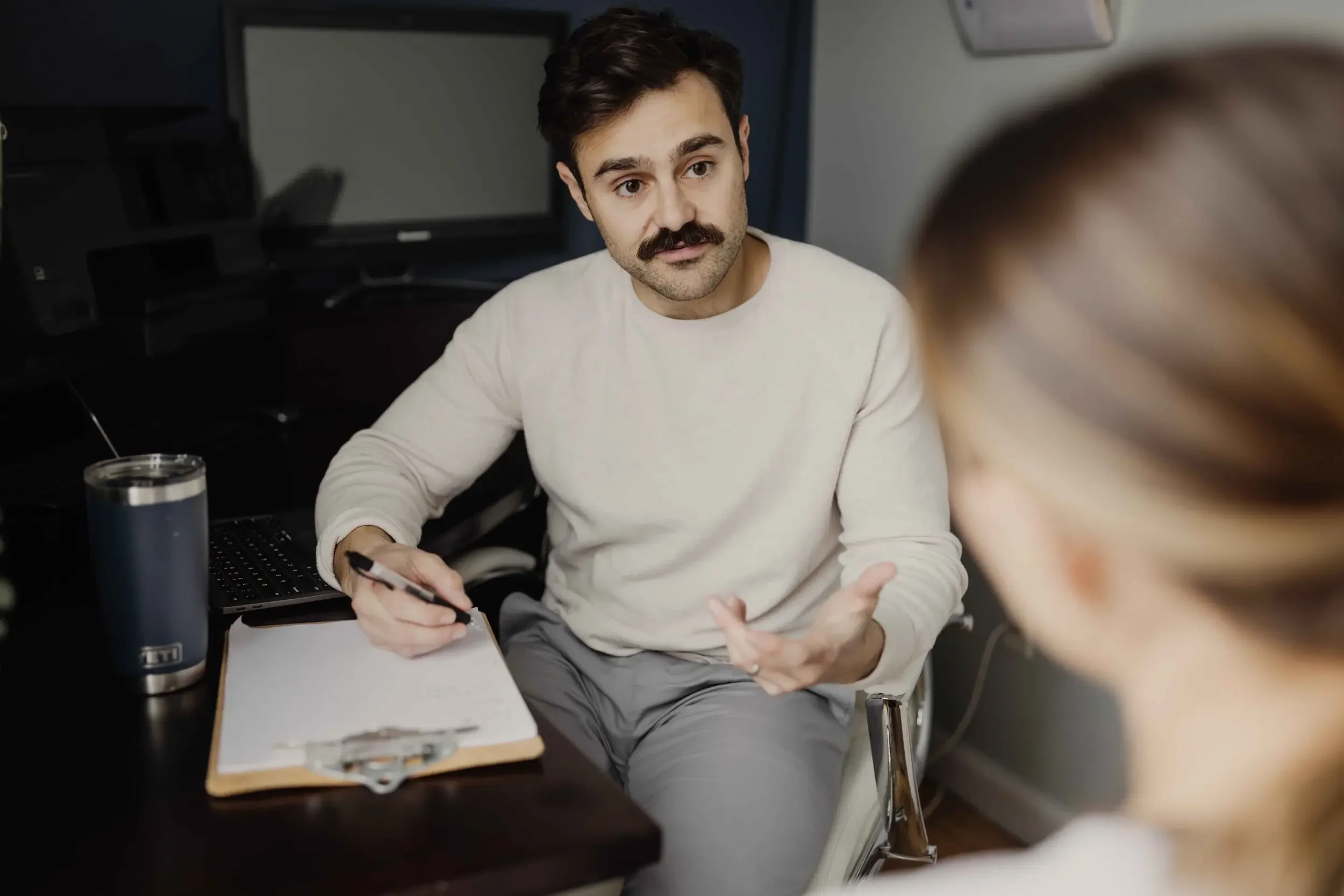 Man with mustache sitting at desk talking to a person
