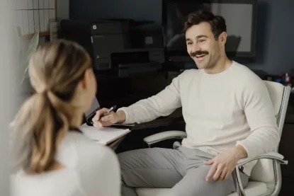 A man with dark hair, a mustache, and light skin smiling while talking to a woman with light skin and brown hair tied back, in an office setting.