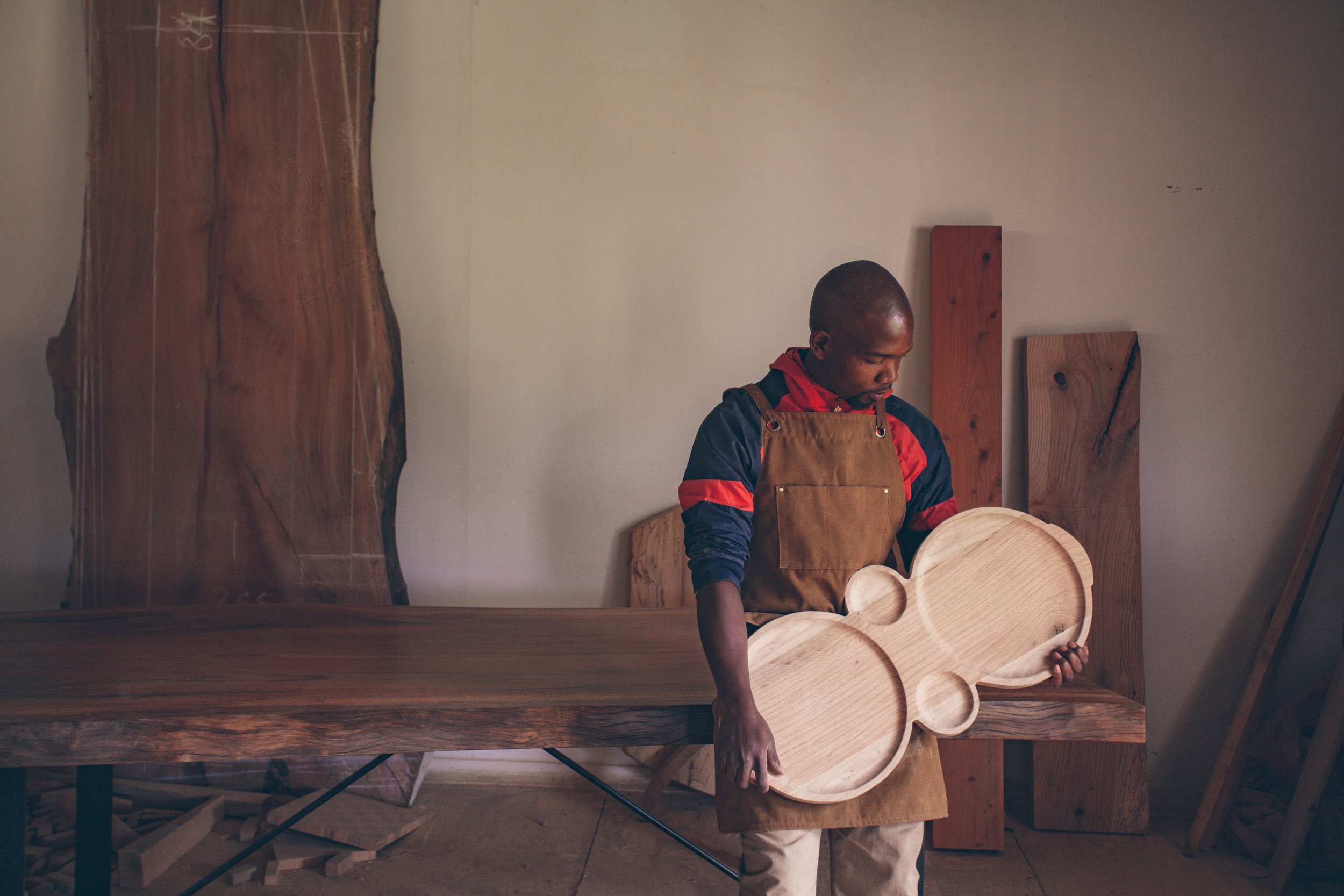 A man in a red and black jacket and a tan apron is holding a carved wooden guitar body in a woodworking shop with wooden slabs and planks in the background.