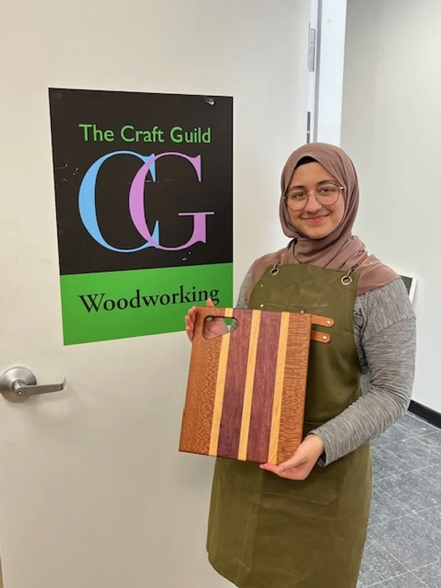 A young woman with glasses and a hijab, smiling, holding a wooden cutting board with striped wood design, standing in front of a sign that reads 'The Craft Guild Woodworking.'