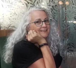 A woman with long curly gray hair, wearing glasses and a black shirt, smiling and resting her chin on her hand indoors.