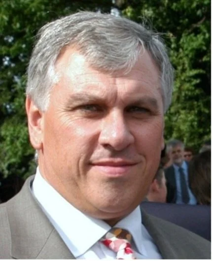 A middle-aged man with gray hair in a suit and tie, outdoors with trees and other people in the background.