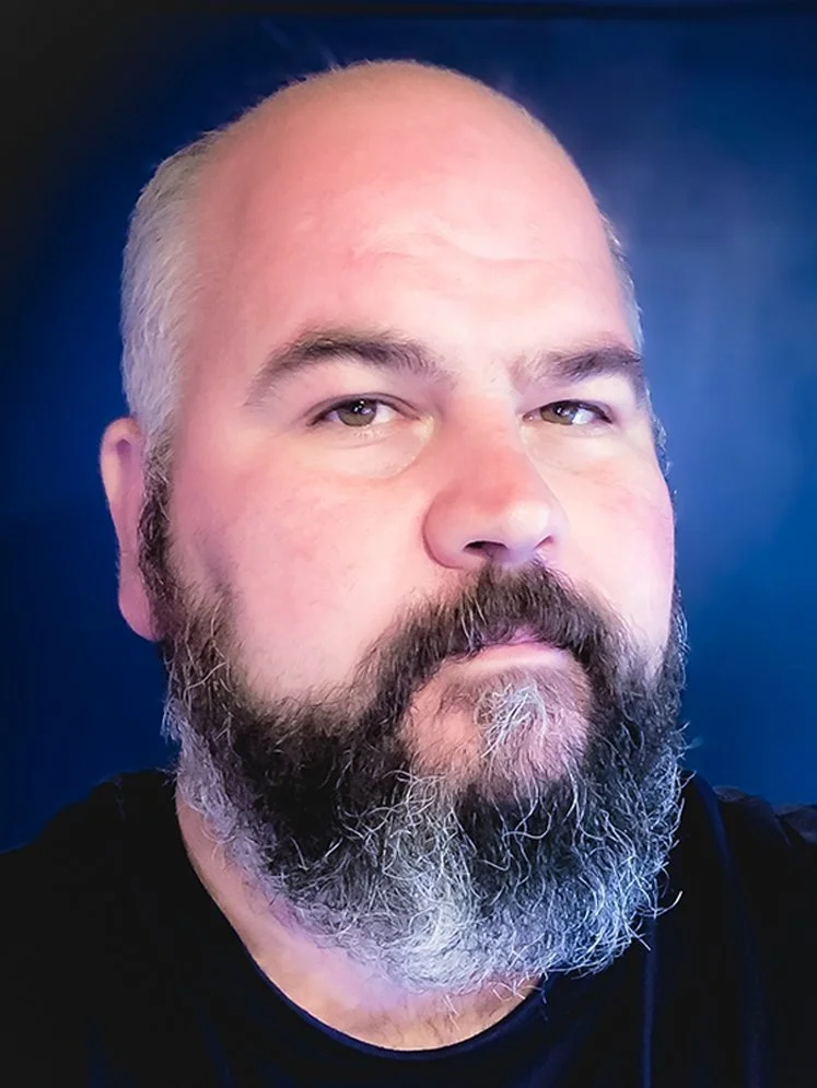 Close-up of a middle-aged man with a beard and mustache, wearing a black shirt, against a dark blue background.