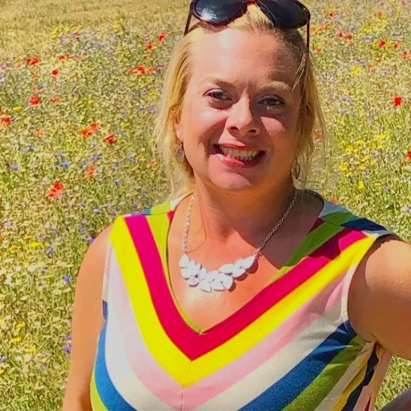 Close-up of a woman wearing dark sunglasses, smiling outdoors at the beach, with people walking in the background.