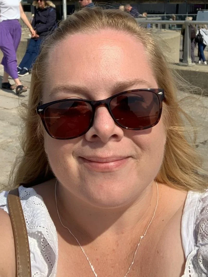 Close-up of a woman wearing dark sunglasses, smiling outdoors at the beach, with people walking in the background.