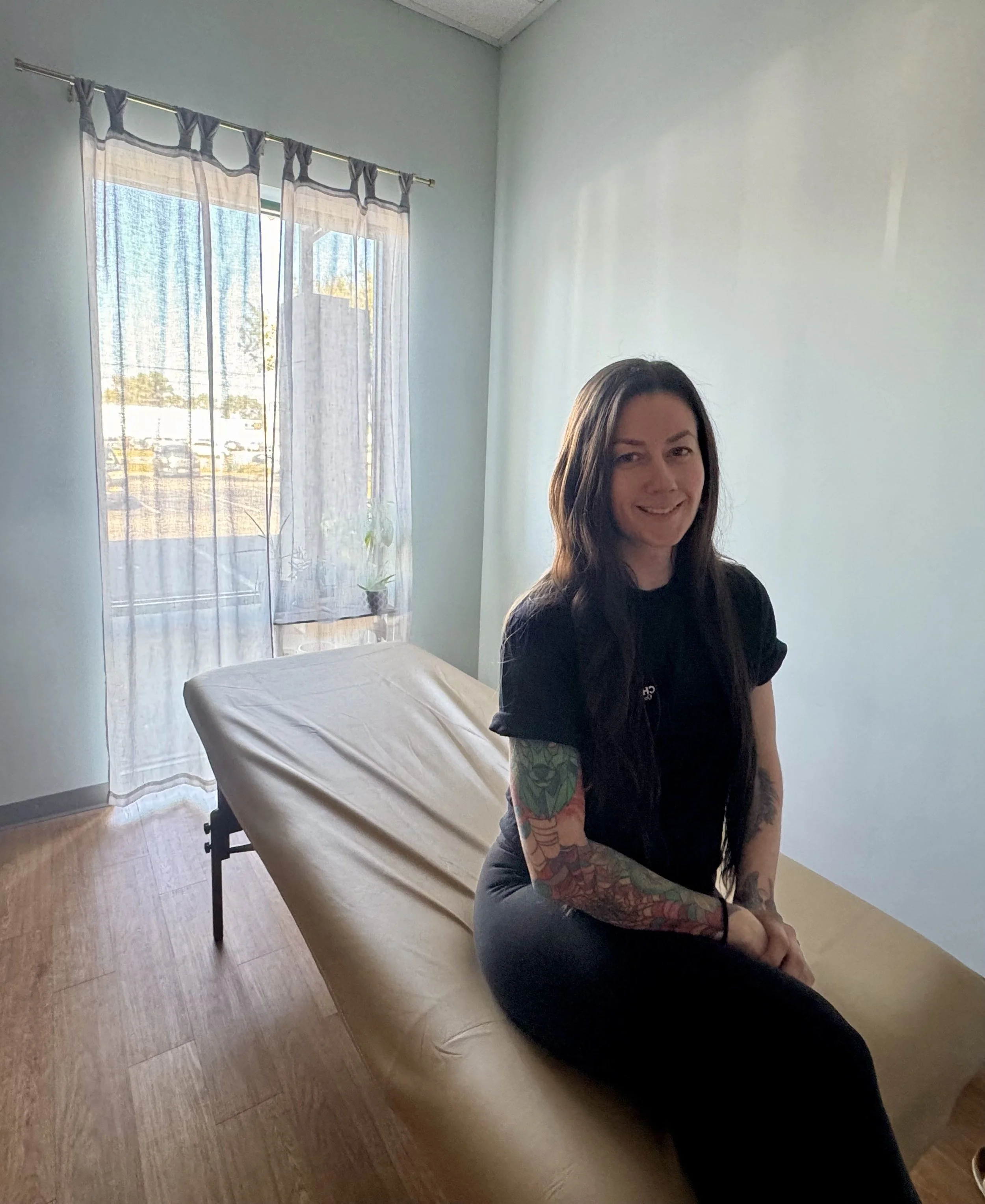 A woman with long dark hair and tattoos on her arms sitting on a massage table in a lightly lit room with a window and sheer curtains.
