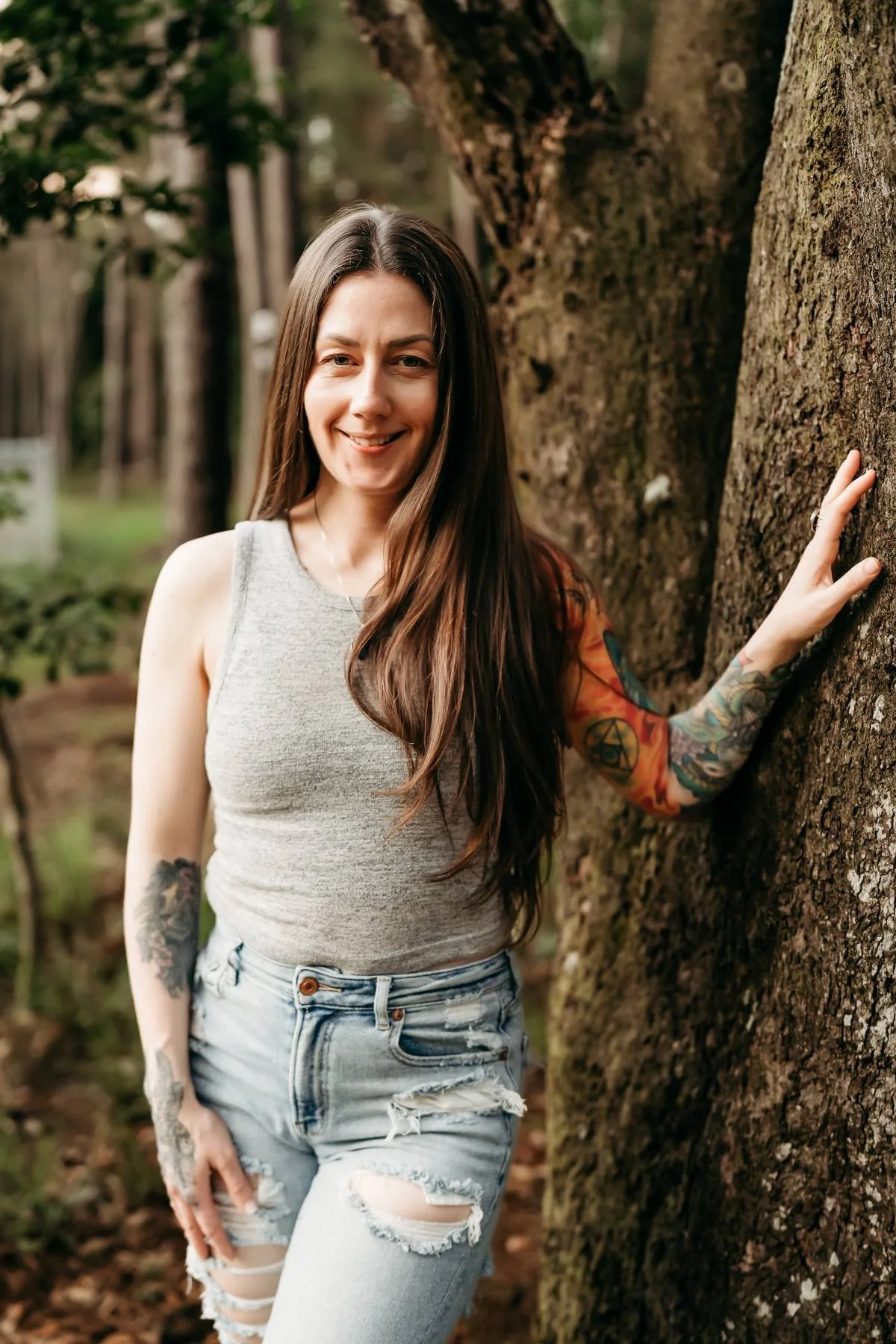 A woman with long dark hair and tattoos, wearing a sleeveless gray top and ripped jeans, standing outdoors next to a tree and smiling.