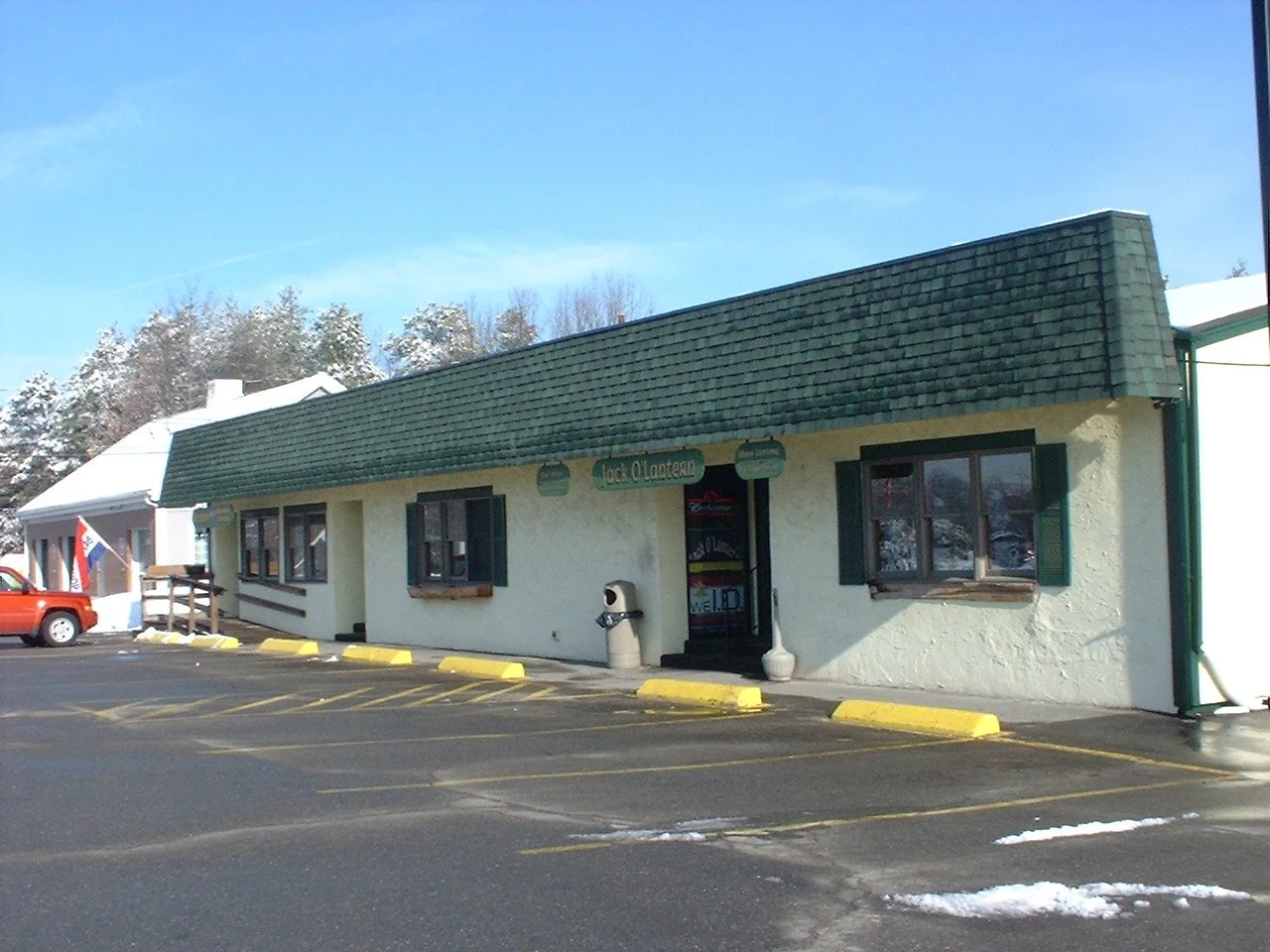 Exterior of a small restaurant called 'Jack O Lantern' with a parking lot in front, a red truck parked on the side, some snow on the ground and trees in the background.