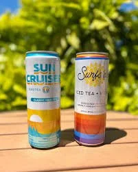Two tall cans of sparkling water and iced tea resting on a wooden surface outdoors with green foliage in the background.