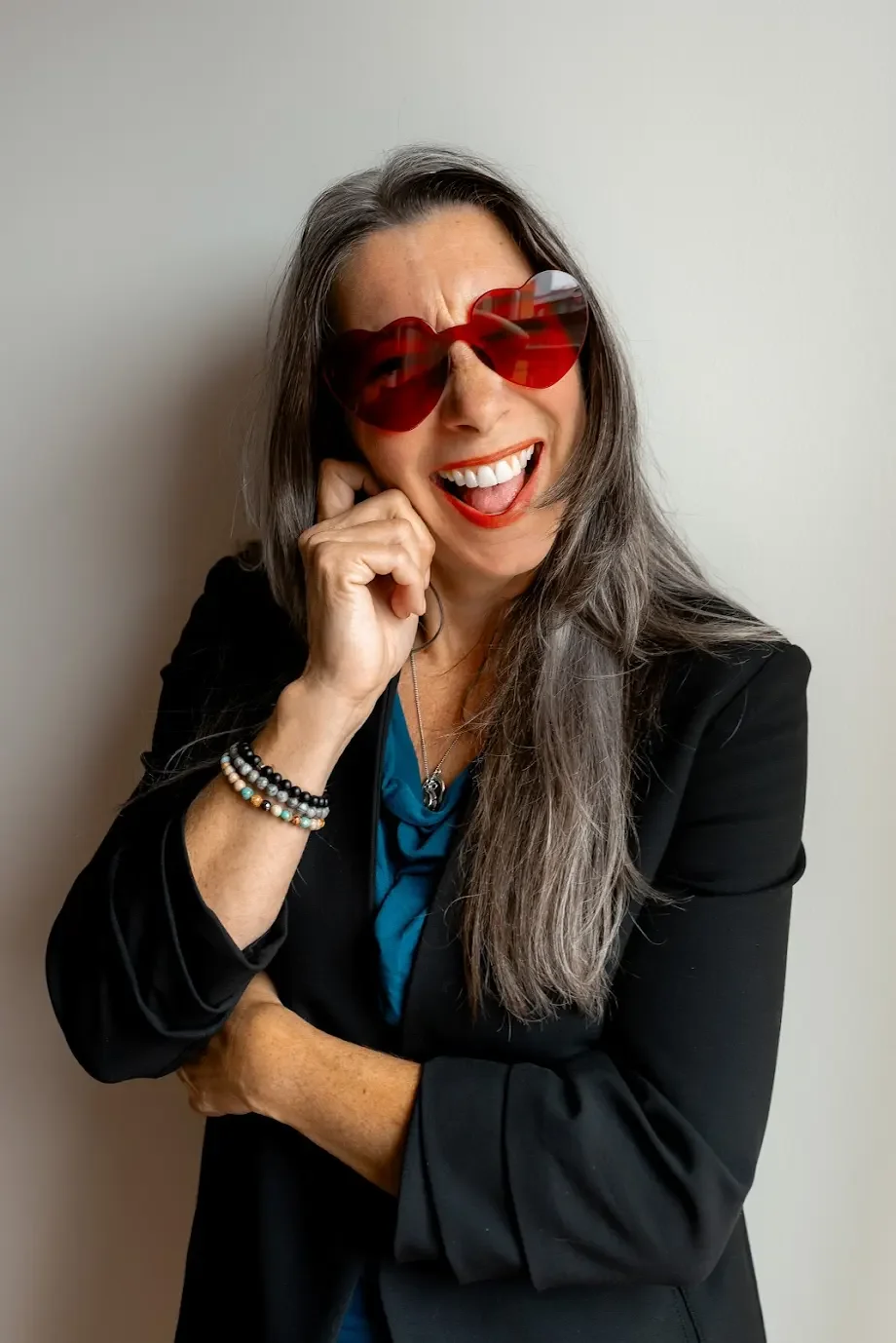A woman with long gray hair wearing red heart-shaped sunglasses, a black blazer, and a blue top, smiling and laughing with her hand near her face against a plain white wall.
