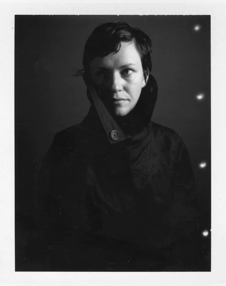 Black and white portrait of a person with short dark hair, wearing a dark jacket with a high collar, against a dark background with small light spots.
