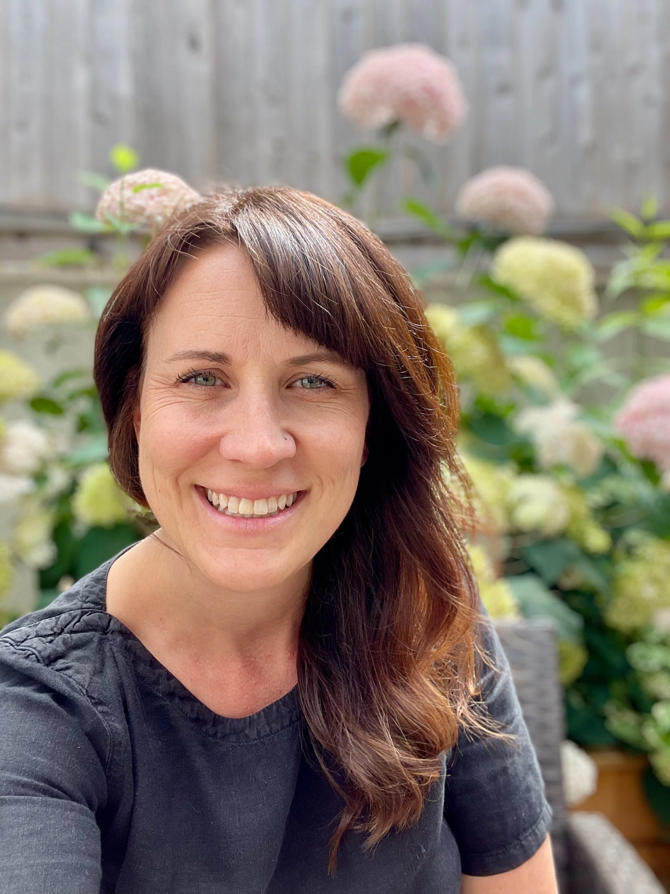 A woman smiling outdoors, with pink and white hydrangea flowers and a wooden fence in the background.
