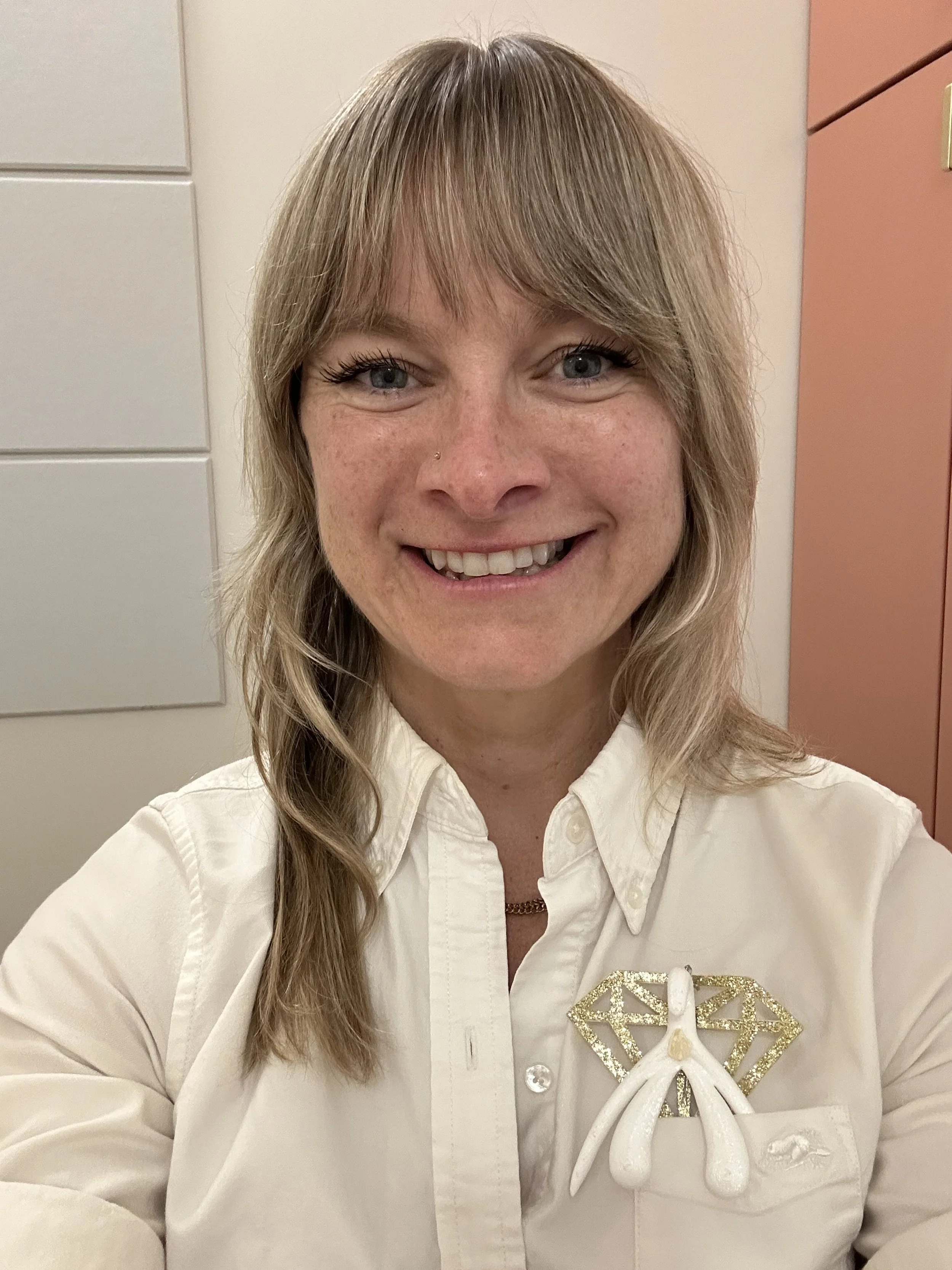 A woman with blonde hair smiling, wearing a white shirt and a decorative butterfly brooch, standing in an indoor setting with cream and orange-colored lockers in the background.