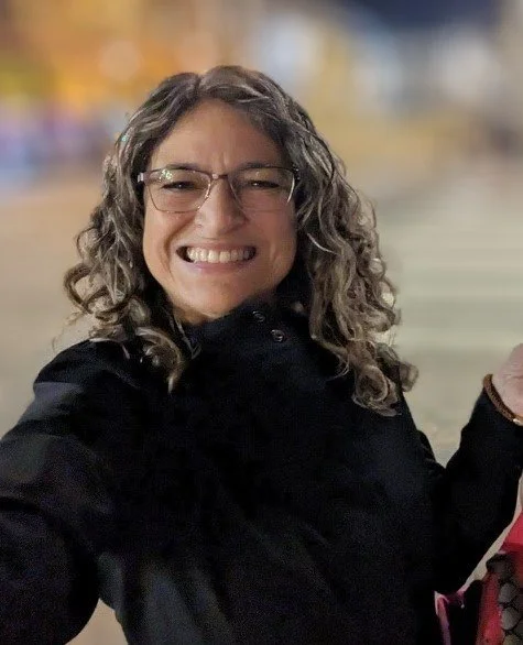 A woman with curly hair and glasses smiling outdoors at night.