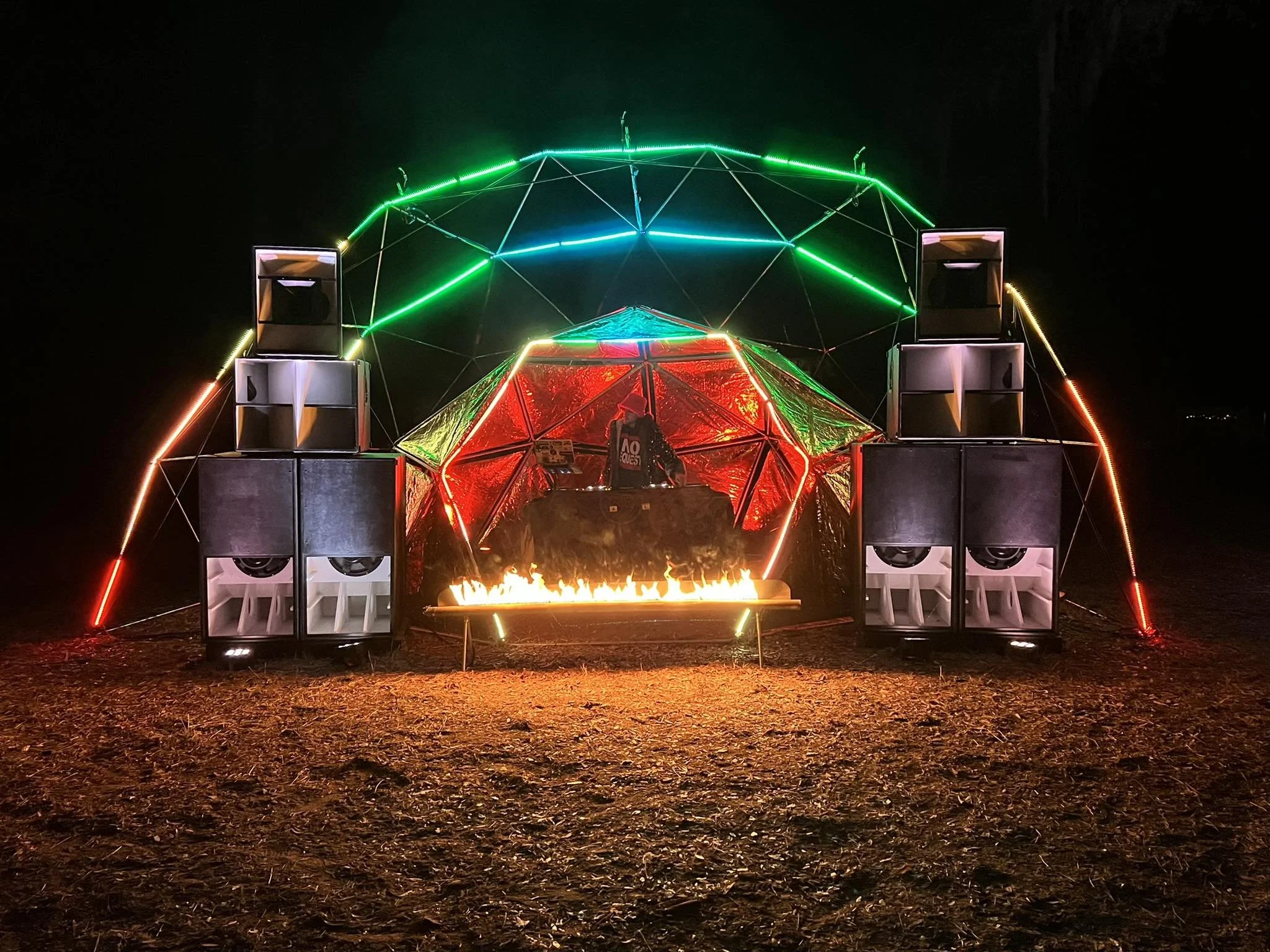 A nighttime scene featuring a geometric dome structure with multicolored neon lights, a DJ playing inside, and a row of speakers in front of the setup.