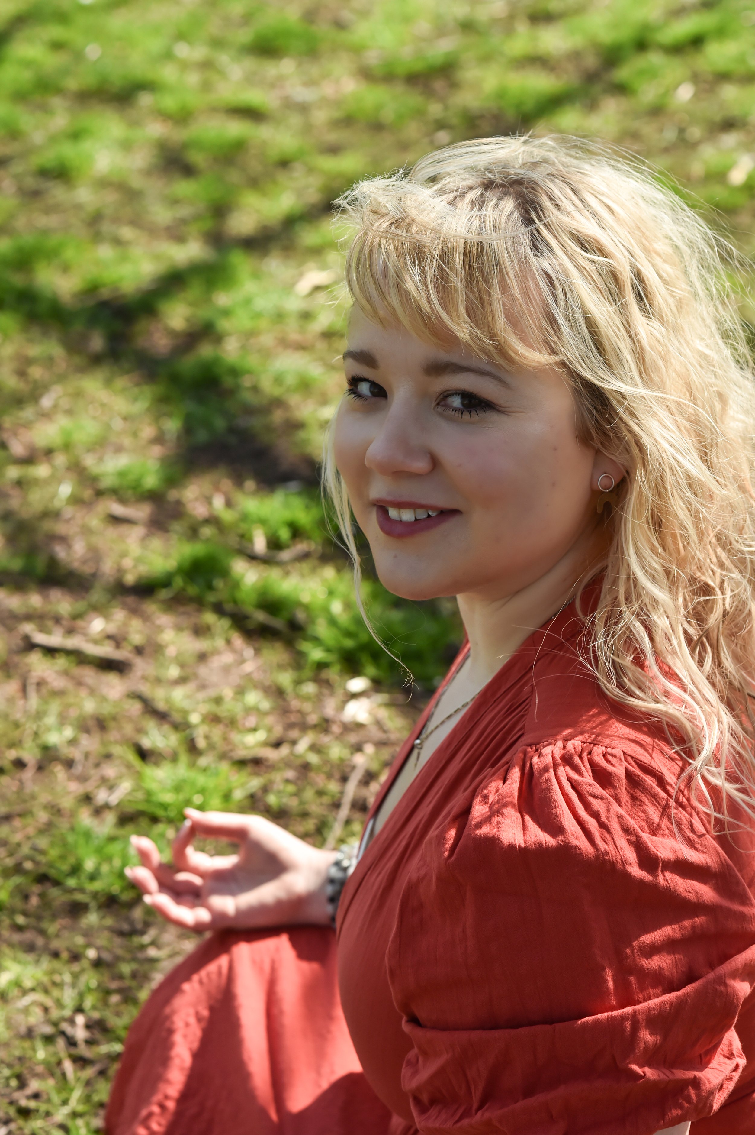 Woman sitting on grass in a red dress, smiling, with sunlight on her face.