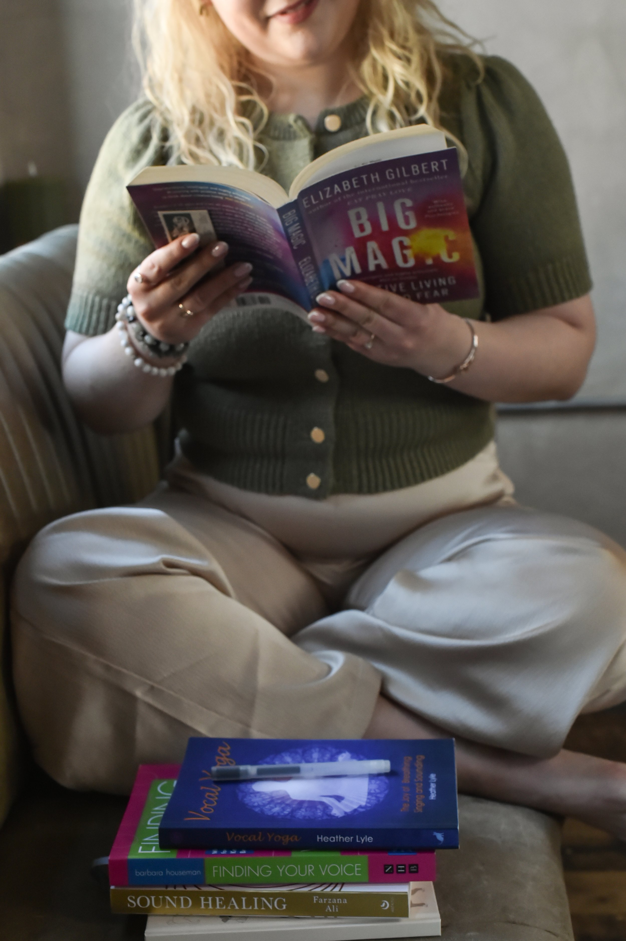 Person reading "Big Magic" by Elizabeth Gilbert, sitting cross-legged on a sofa with a stack of books including "Vocal Yoga," "Finding Your Voice," and "Sound Healing."