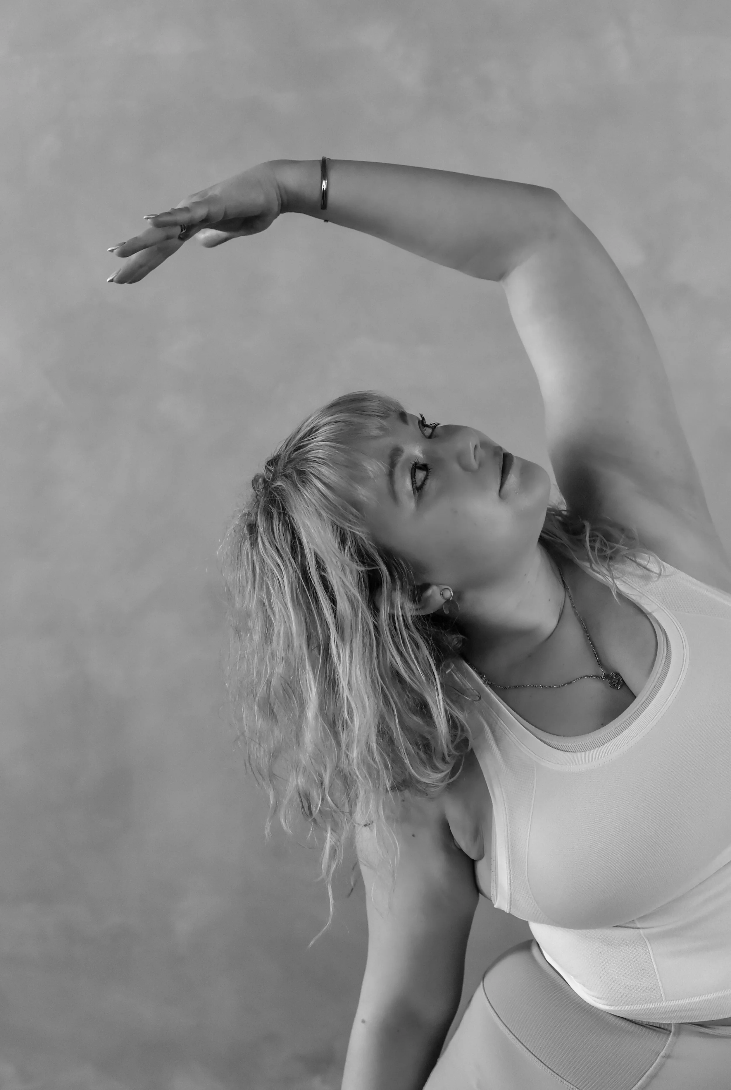 A woman with curly blonde hair performing a yoga pose, with her arm stretched above her head, against a plain background, in black and white.