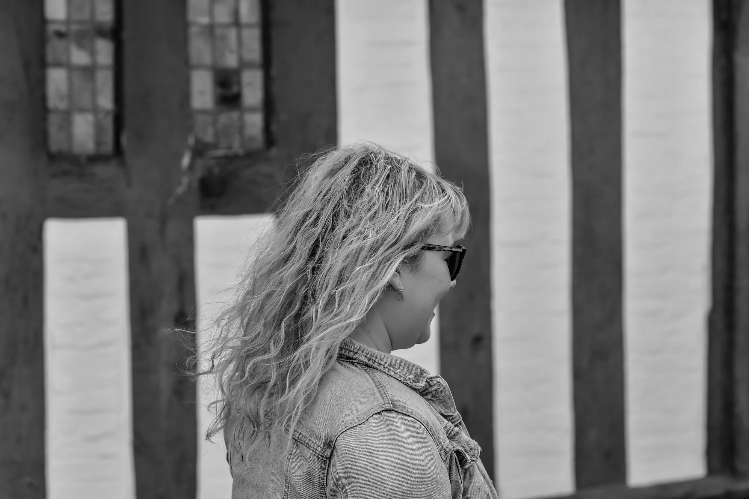 A woman with wavy blonde hair wearing sunglasses and a denim jacket stands in front of a wooden fence, looking downward.