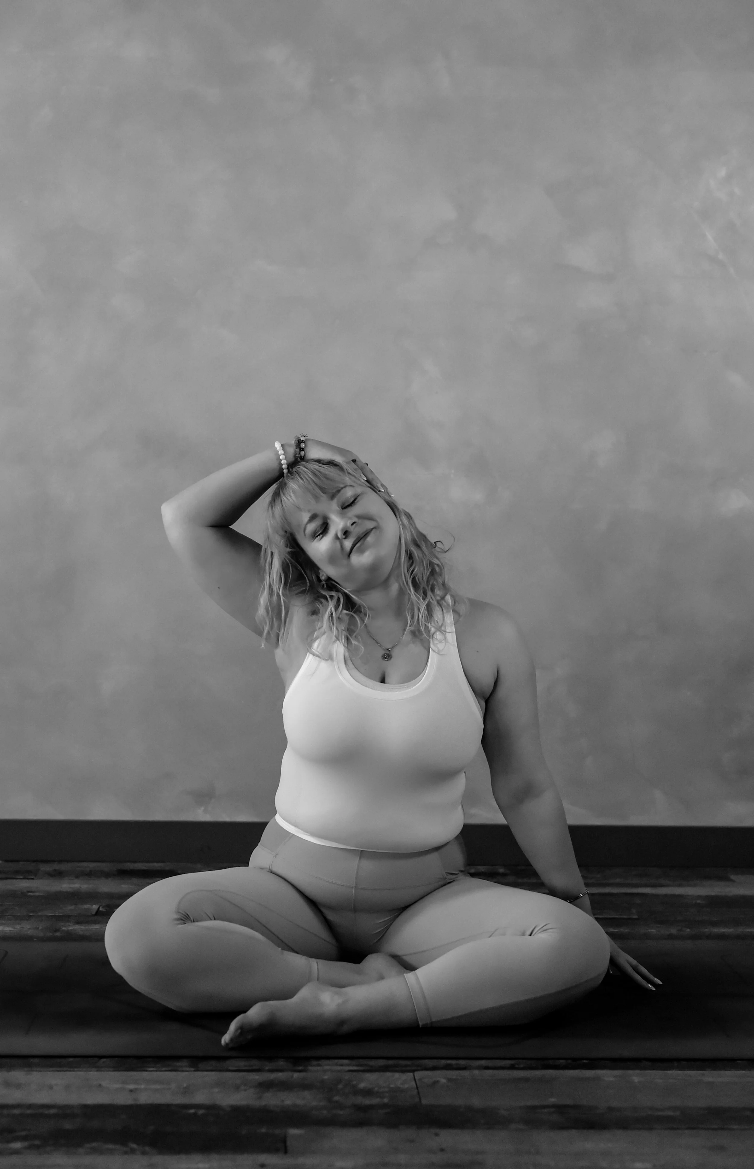 A woman practicing yoga in a seated cross-legged pose, smiling with her eyes closed, with her right hand resting on her head and her left hand on the floor. She is wearing a tank top and leggings, and is sitting on a yoga mat against a plain wall.