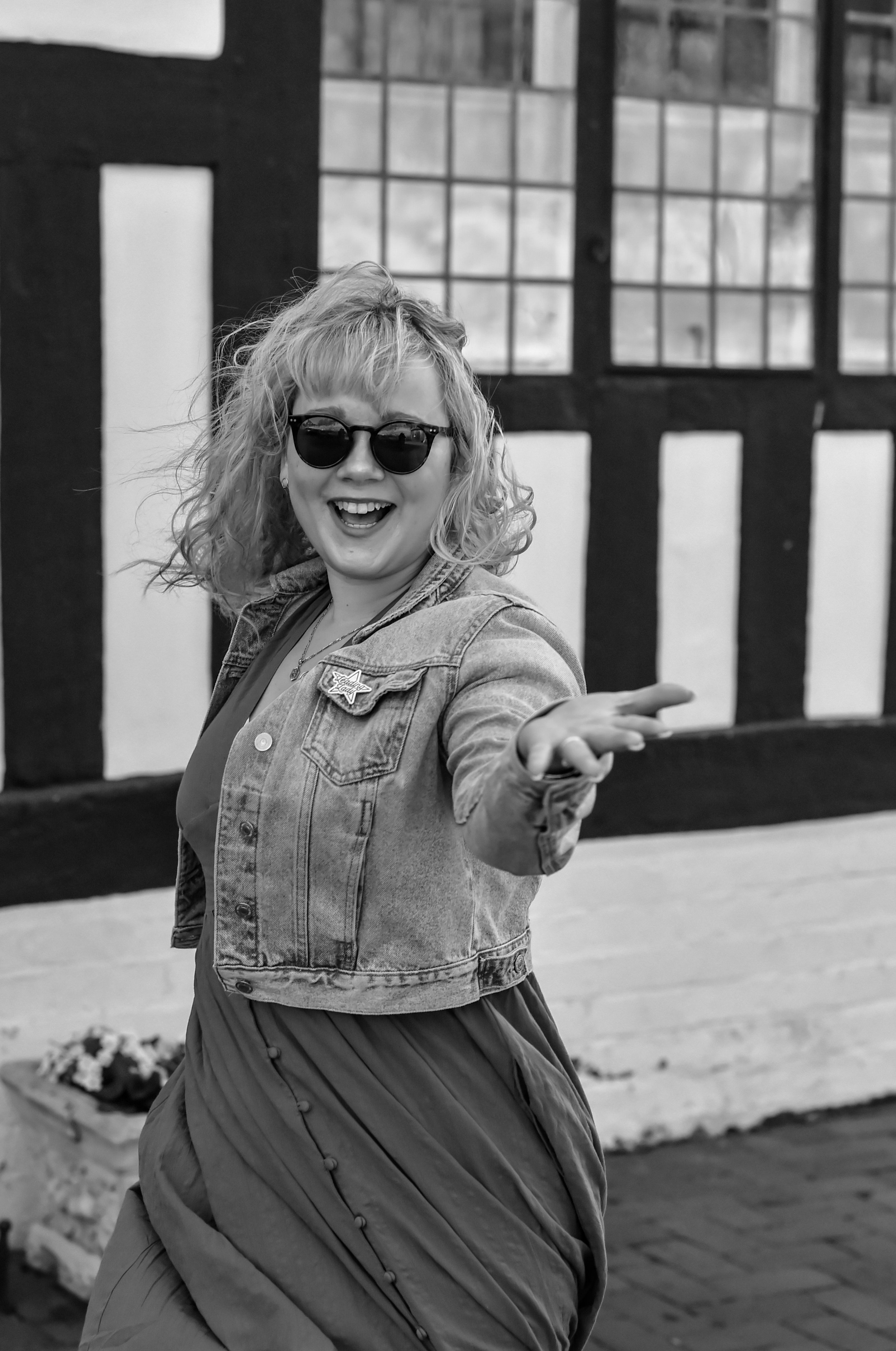 Black and white photo of a woman wearing sunglasses, a denim jacket, and a dress, smiling and reaching out with her hand in front of a Tudor-style building.