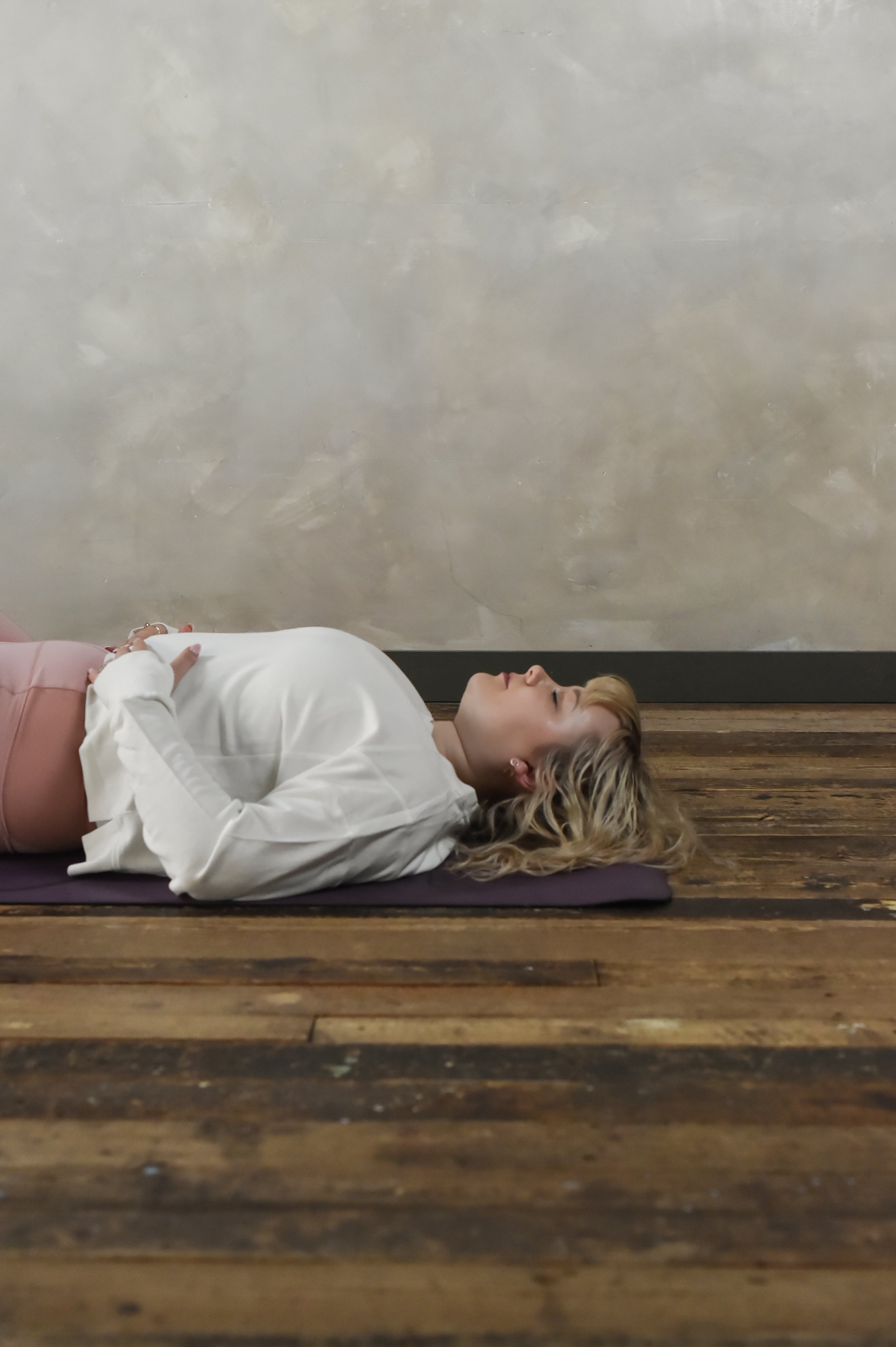 Woman lying on a yoga mat on a wooden floor, wearing a white long sleeve and pink pants with hands on her stomach.