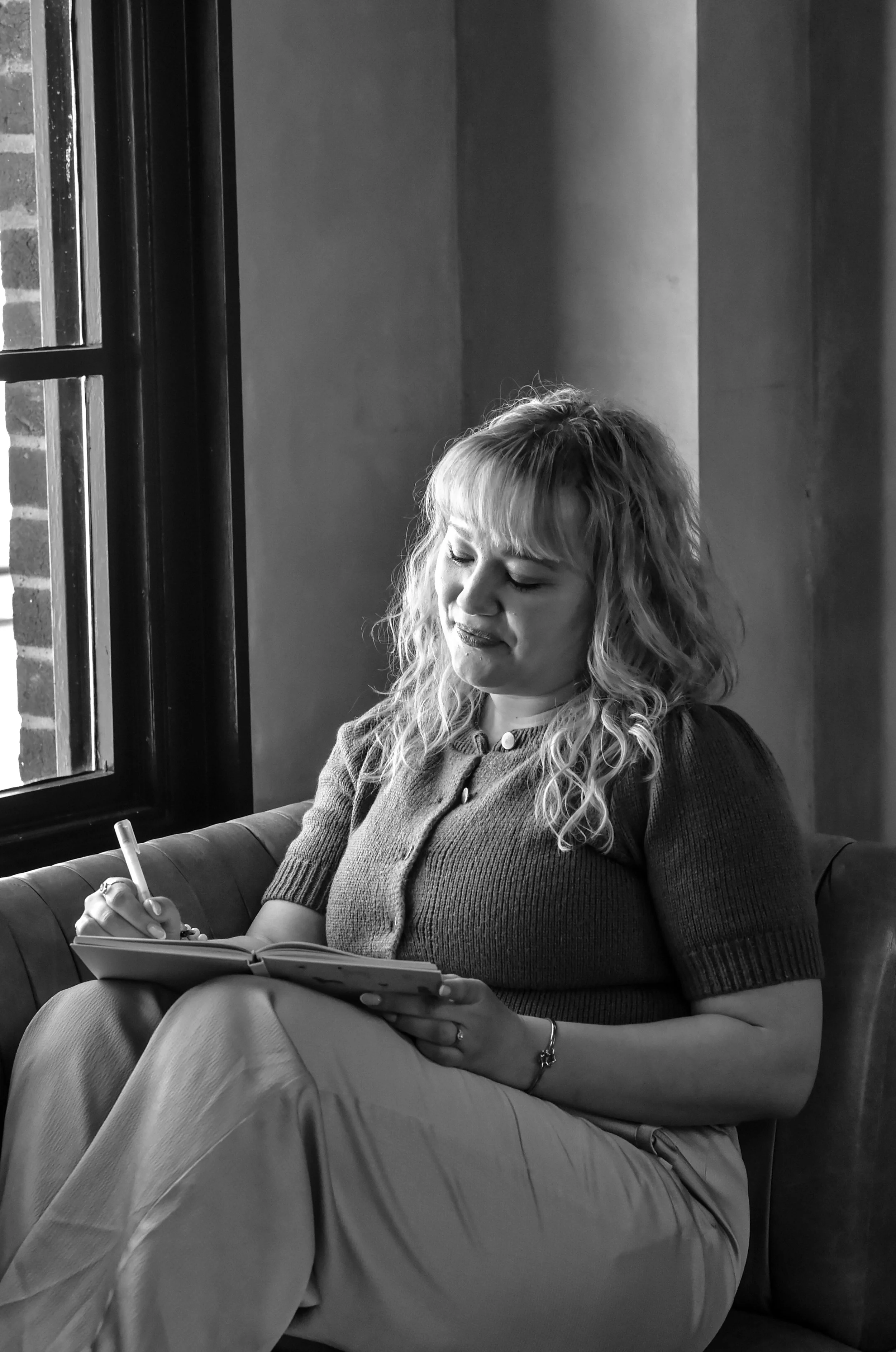 A woman with curly hair and a top with buttons, sitting on a cushioned bench near a window, writing in a notepad.