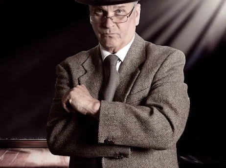 Jeremy Todd acting photo, from "Home at Seven" play. A man wearing glasses, a gray suit, and a tie, with his arms crossed, in a dimly lit setting.