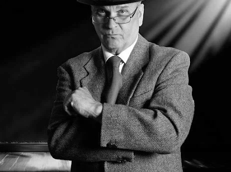 Jeremy Todd acting photo, in the "Home at Seven" play. Black and white photo of a man wearing glasses, a suit, and a hat, with his arms crossed and looking at the camera.