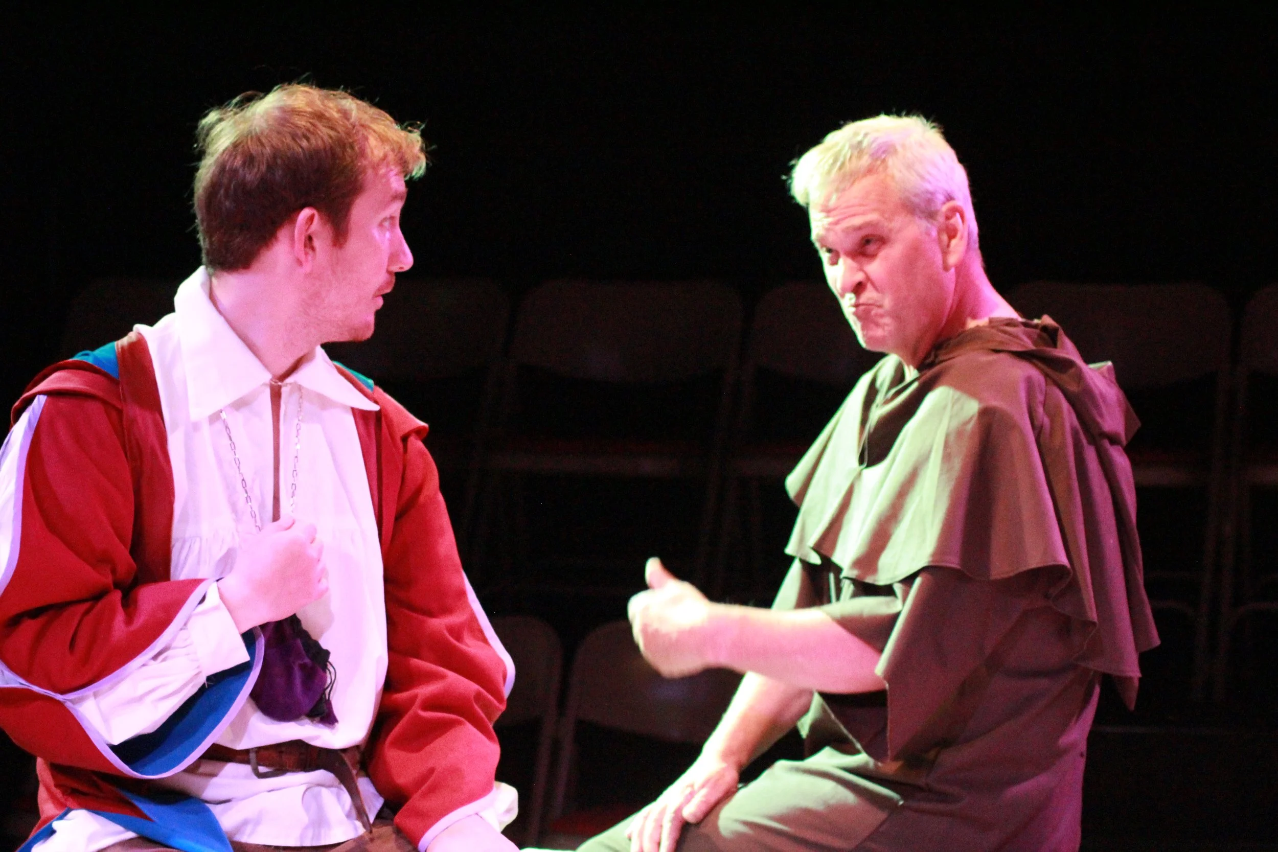 Jeremy Todd acting photo. Two actors on stage in a theatrical scene, one young man wearing a colorful jacket and the other older man with grey hair wearing a brown cape, engaged in a conversation.