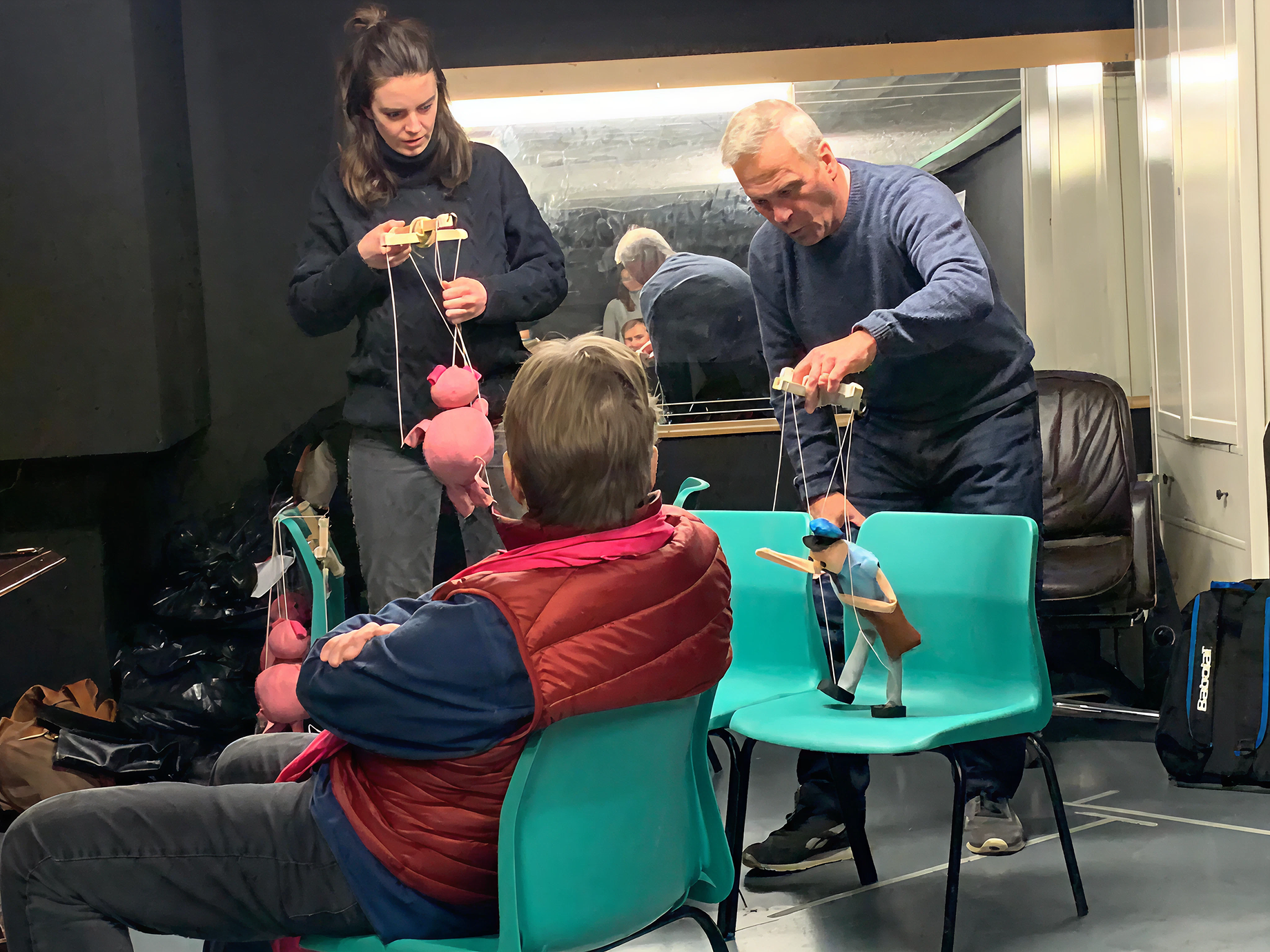Two puppeteers, a woman and a man, perform marionette puppets for a seated audience member in a room with black walls and a mirror. The woman holds pink pig puppets, while the man manipulates a puppet dressed in blue. The audience member has his hands crossed and wears a red vest.