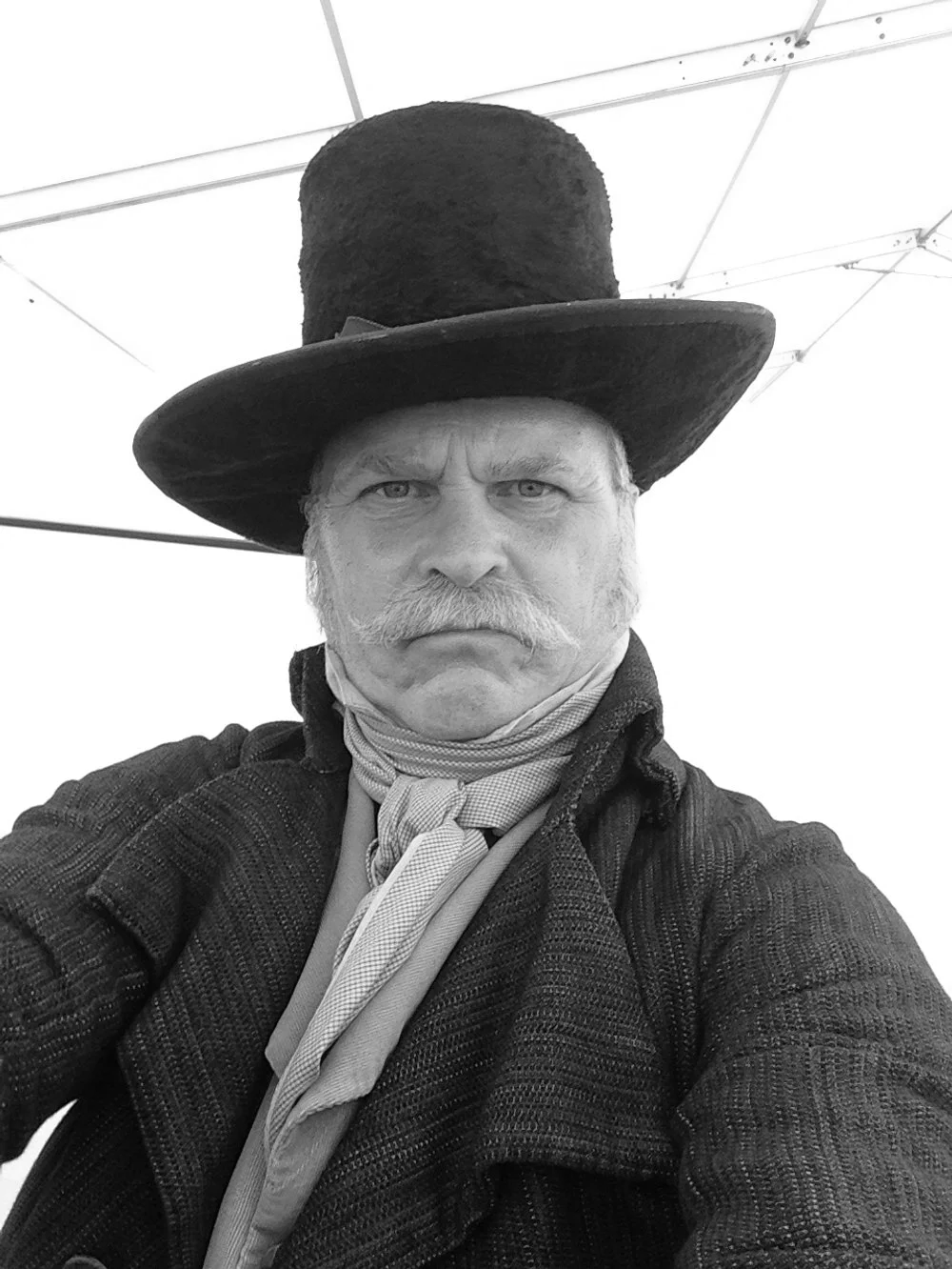 Jeremy Todd acting photo, as actor dressed up at Edinburgh FRINGE festival. A man with a stern expression, dressed in historical costume, wearing a tall top hat, a scarf, and a jacket, in front of a tent or canopy.