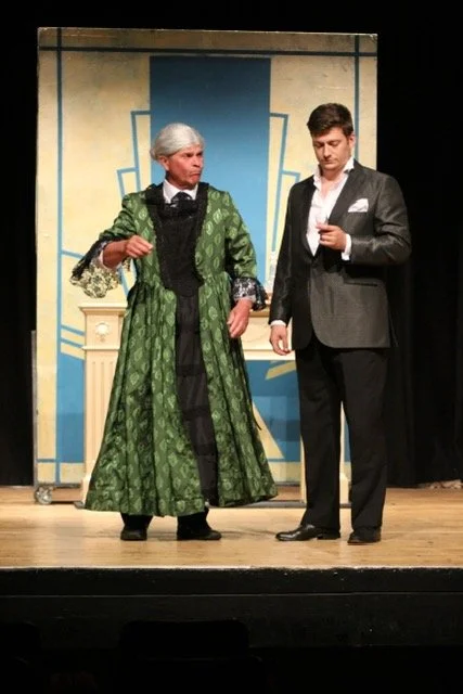 Jeremy Todd acting photo. Two actors on a stage, one dressed in a green vintage dress with a gray wig and the other in a modern suit, engaging in a theatrical performance.
